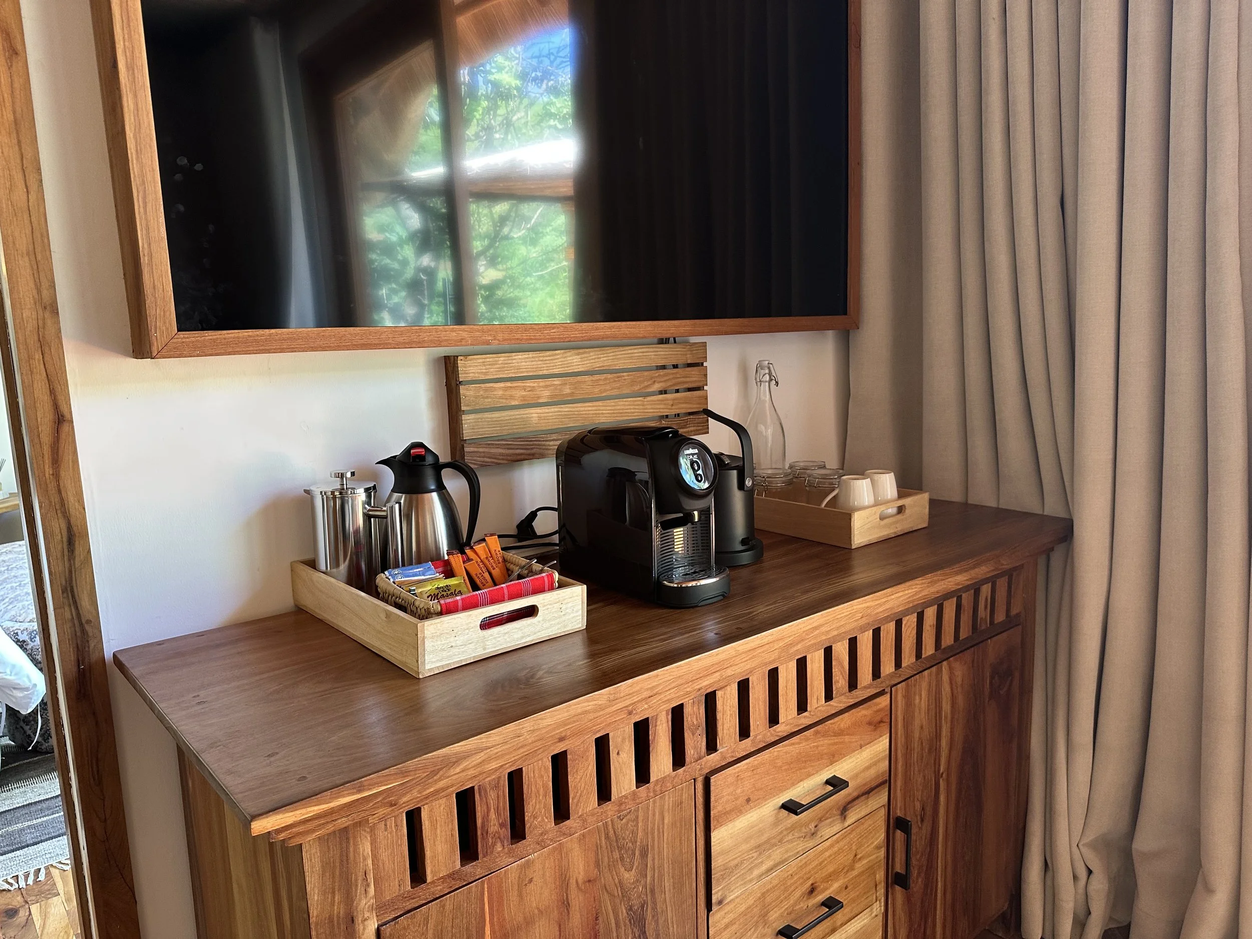 Coffee station on a wooden cabinet with a black coffee maker, a water kettle, a tray with cups, and snack packets, with a flat-screen TV above and cream curtains to the right.
