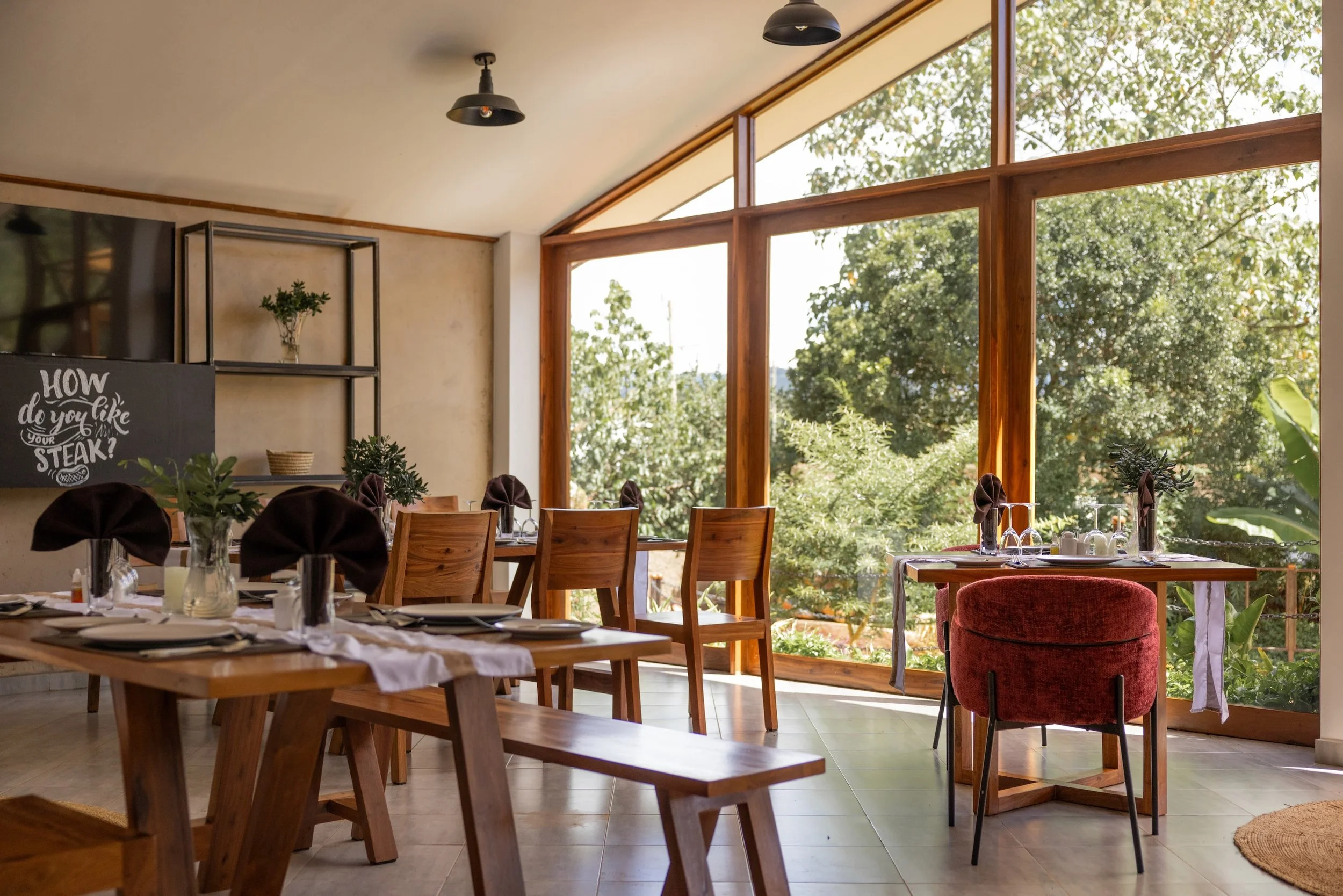 Sunlit restaurant with wooden tables and chairs, set with black cloth napkins, glassware, and tableware, overlooking lush green trees through large floor-to-ceiling windows.