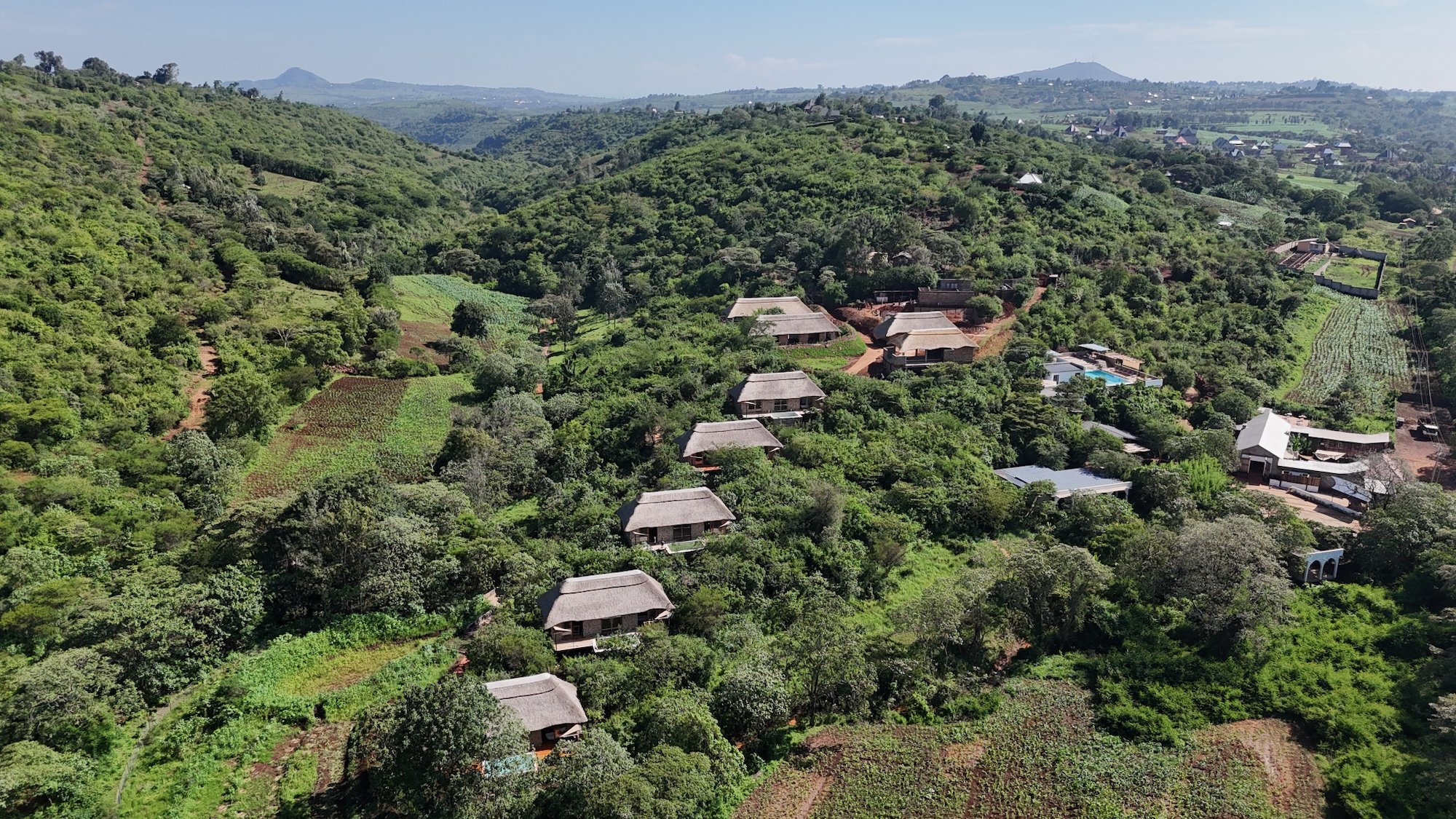 Aerial view of a lush green landscape with hills, small houses with thatched roofs, and cultivated farmland, including trees, crops, and a swimming pool.