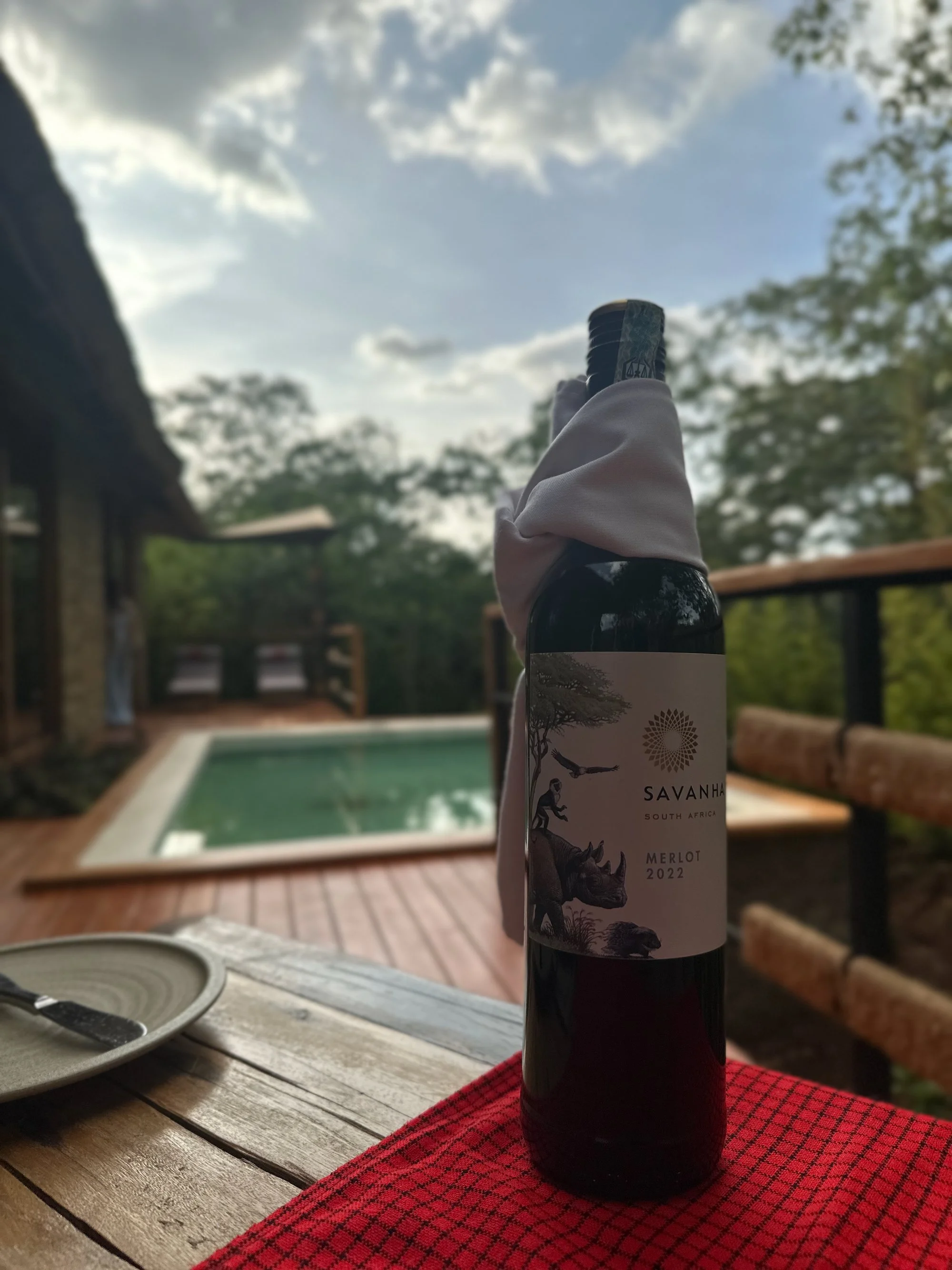 A bottle of Savanna South African Merlot 2022 wine with a white napkin around the neck, standing on a wooden table with a red cloth, with a pool and trees in the background on a partly cloudy day.