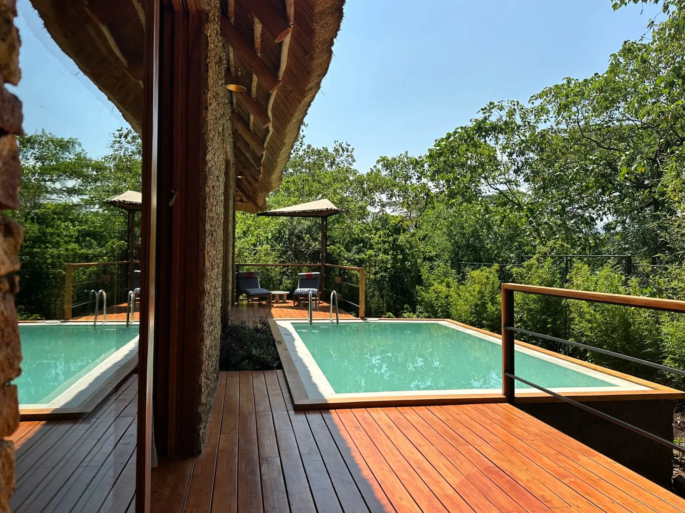 Outdoor pool area with wooden deck, lounge chairs, and a small covered seating area, surrounded by green trees and blue sky.