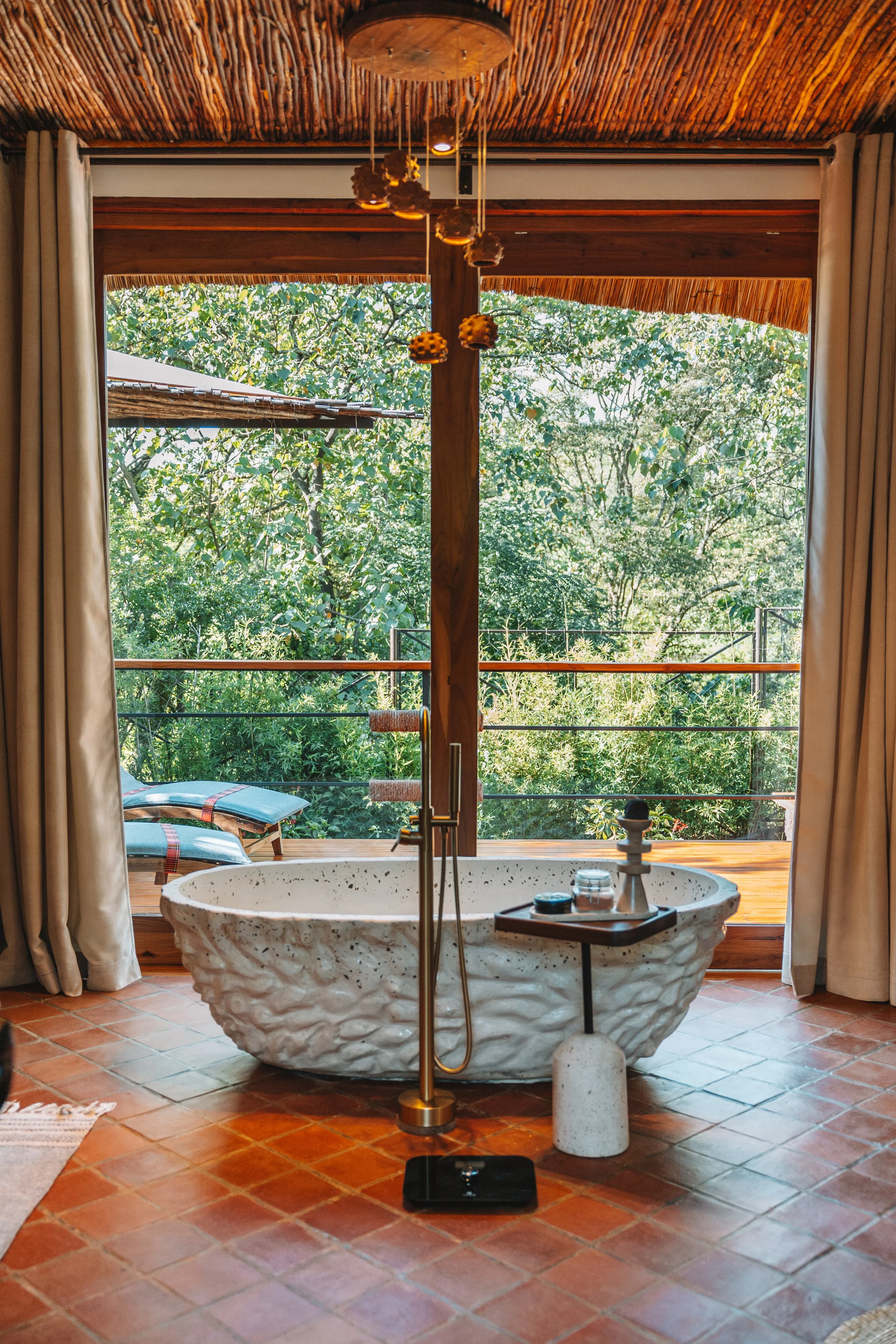 Luxurious bathroom with a freestanding stone bathtub in front of large windows overlooking greenery, with a wooden ceiling and terracotta tile floor.