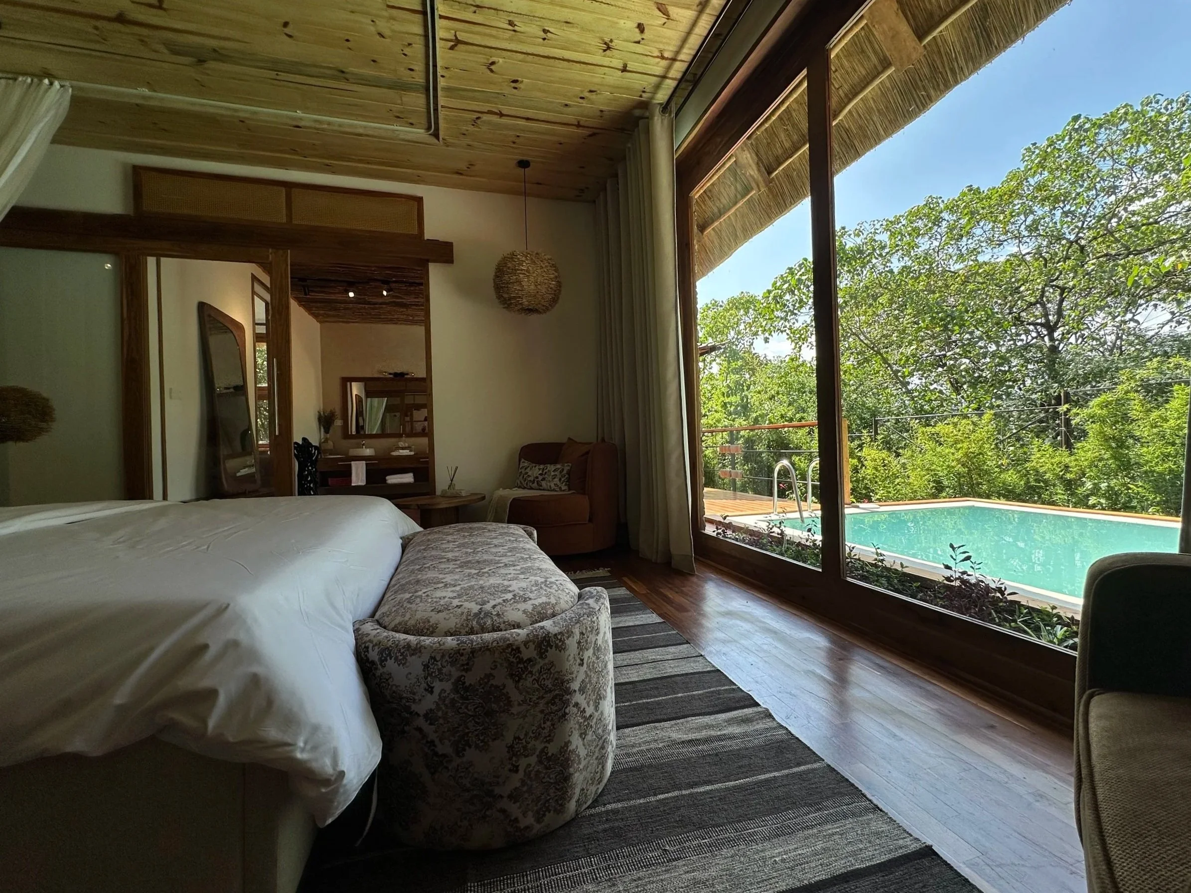 A room with a large glass sliding door overlooking a swimming pool and trees outside, interior with a bed, a bench at the foot of the bed, a chair, and a small table.