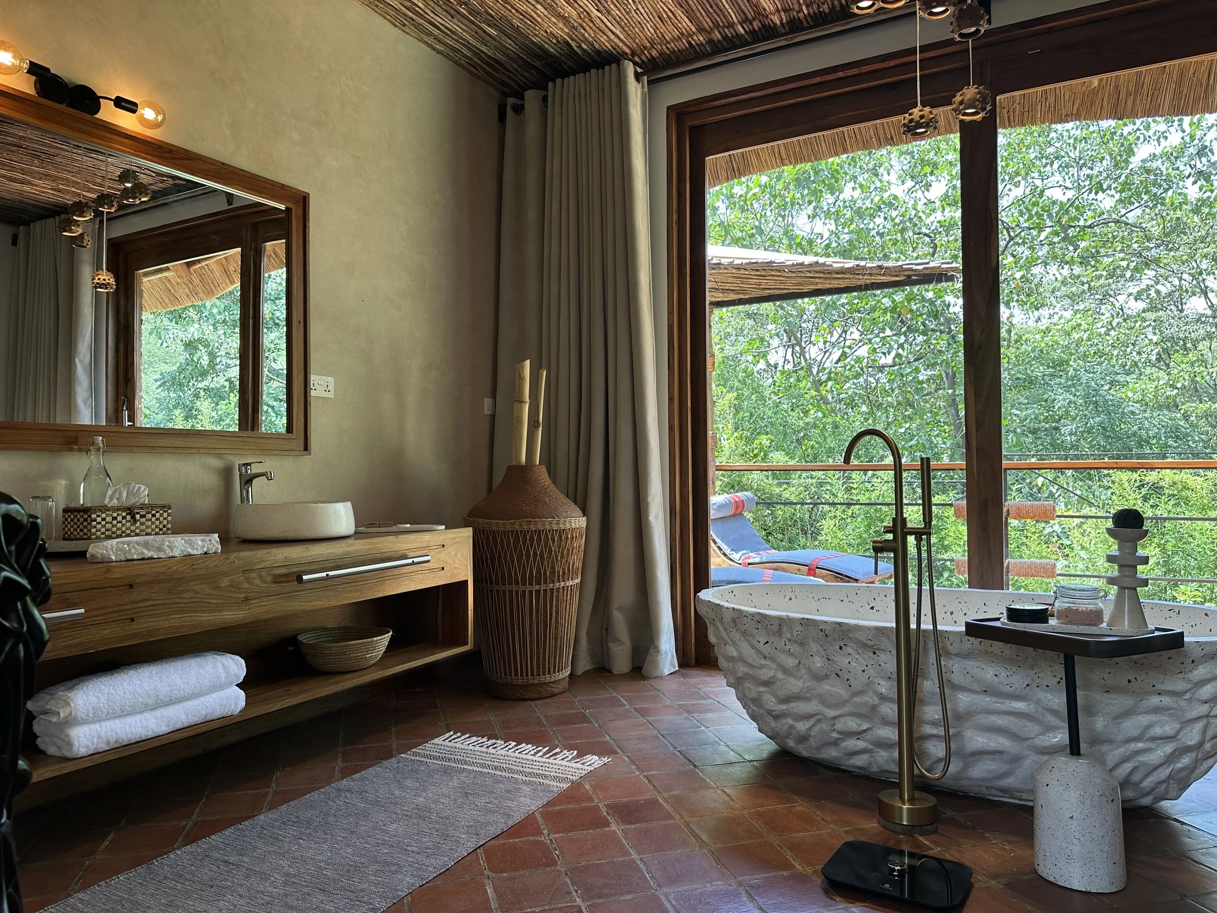 A bathroom with a rustic, natural design featuring a large stone bathtub, a wooden vanity with a sink, a large mirror, a wicker basket with plants, beige curtains, and a view of lush green trees outside through glass doors.
