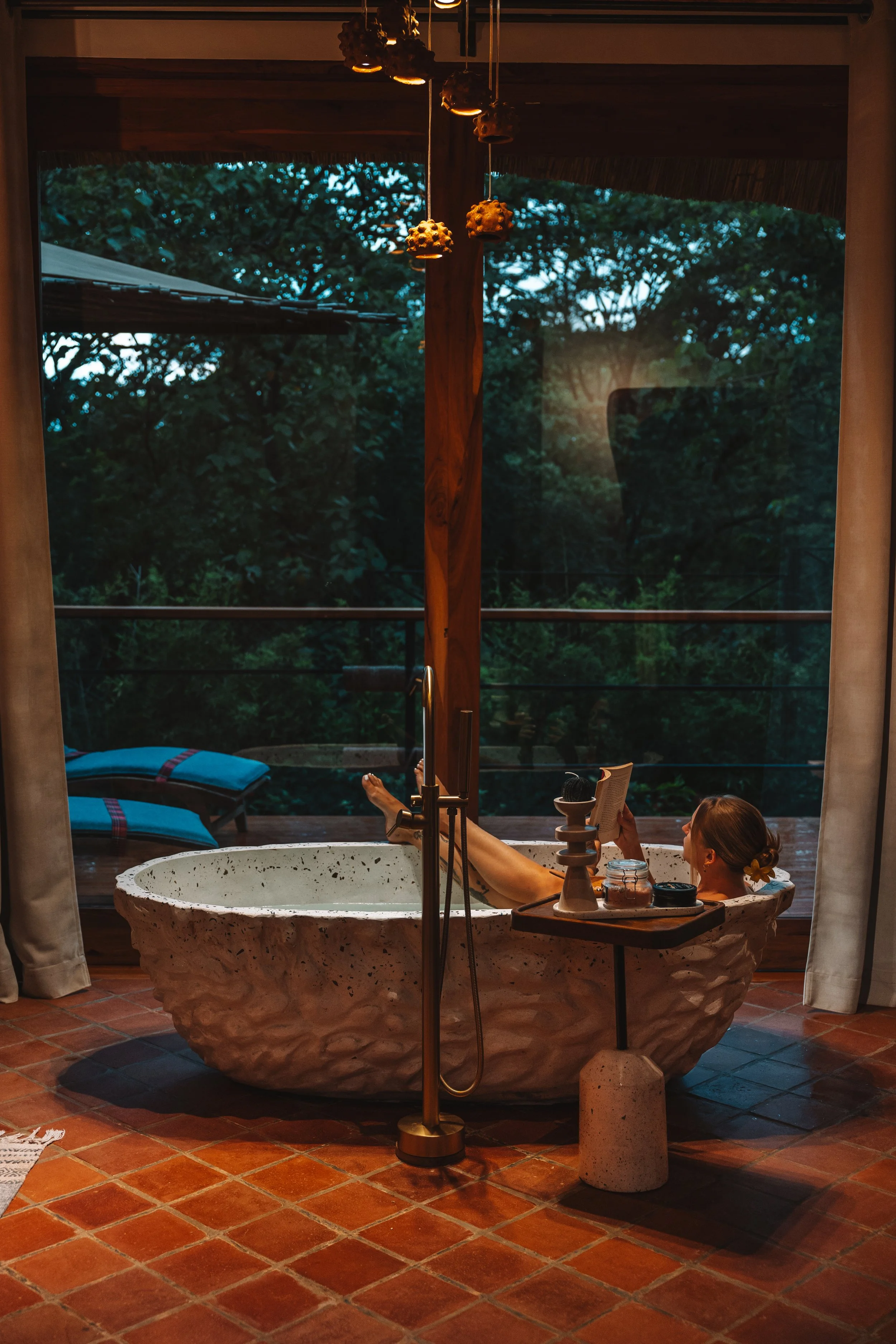A woman relaxing in a rustic stone bathtub on a porch, reading a book at dusk, with a view of trees outside.