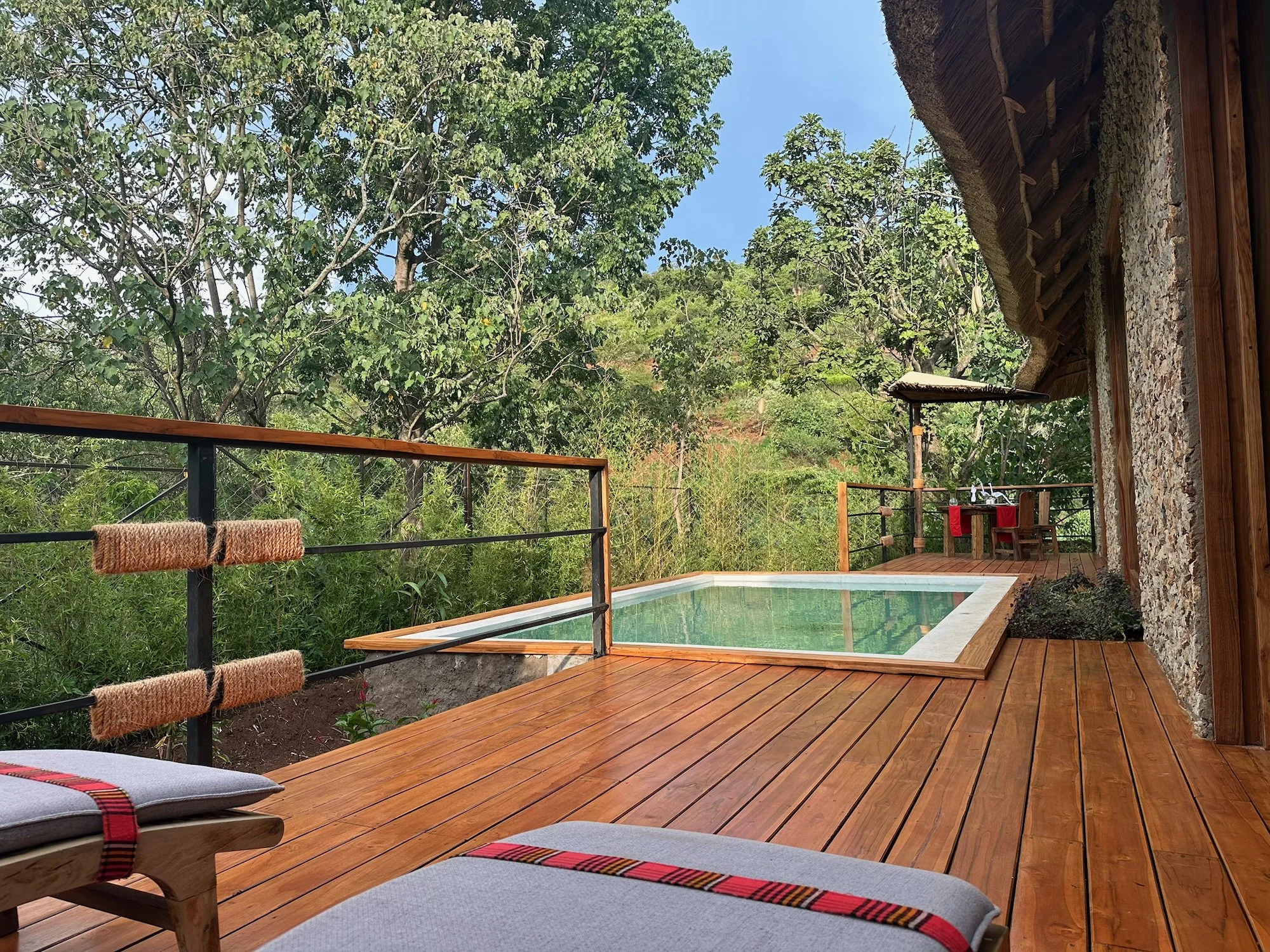 Wooden deck with a small pool, two lounge chairs with cushions, a railing, lush green trees, and a small table with chairs, in a natural outdoor setting.