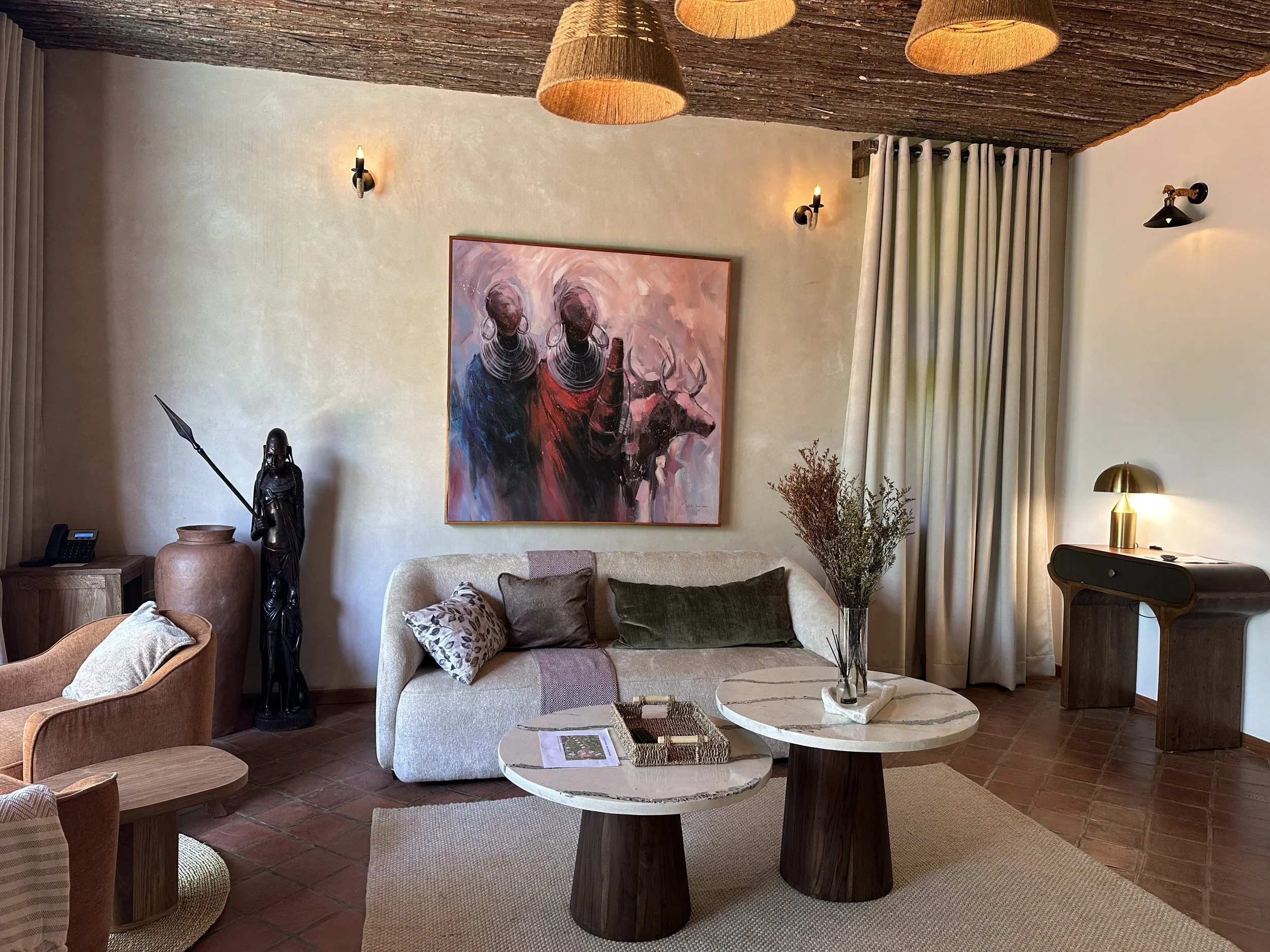 A living room with a beige sofa, decorative pillows, two round marble-top coffee tables, a large painting of tribal figures, a black warrior statue, a large vase, and various lighting fixtures including wall sconces and pendant lights, with curtains covering a window.