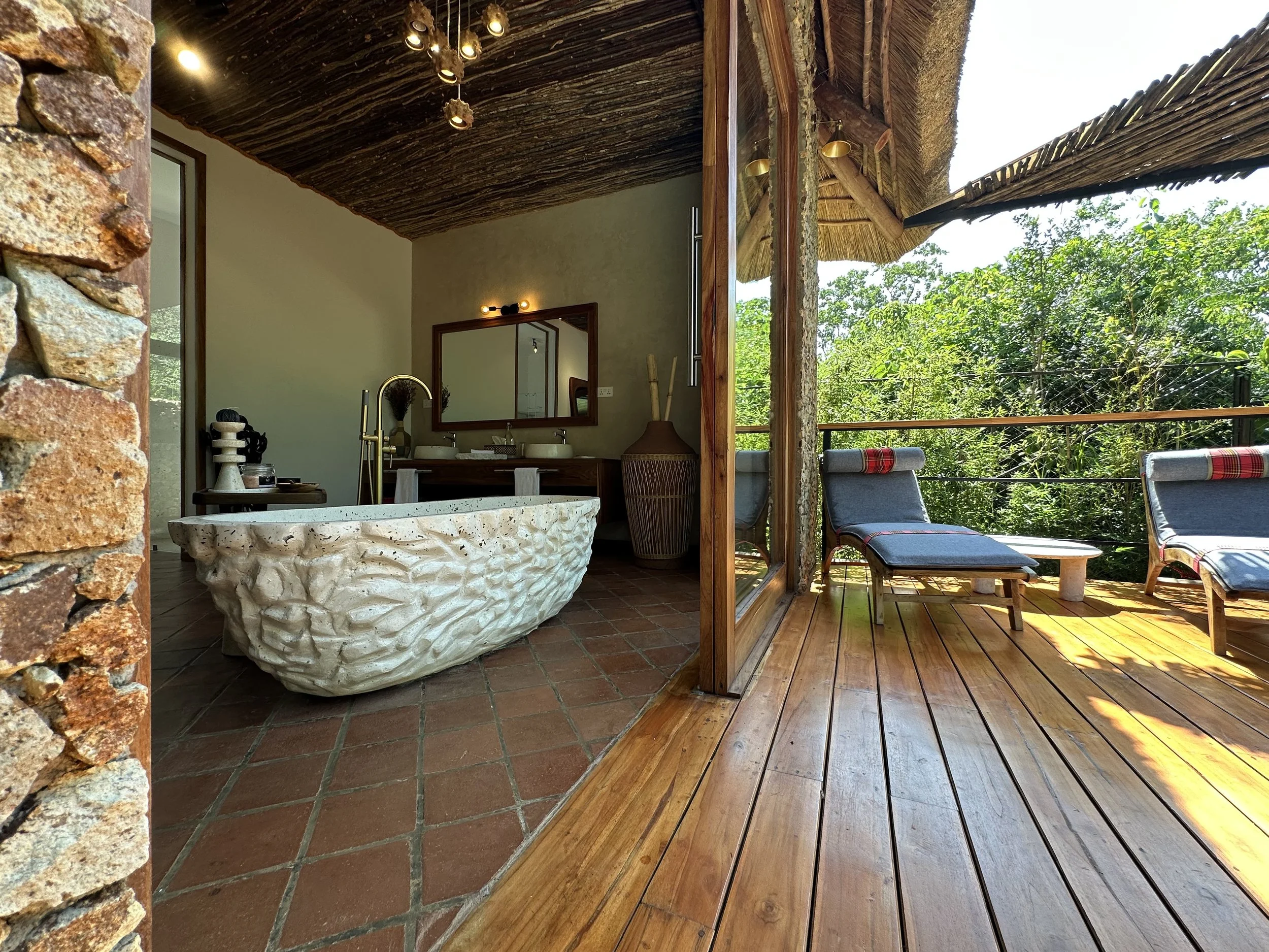 Bathroom with a stone bathtub, large mirror, and double sinks, opening to an outdoor wooden deck with lounge chairs and surrounding trees.