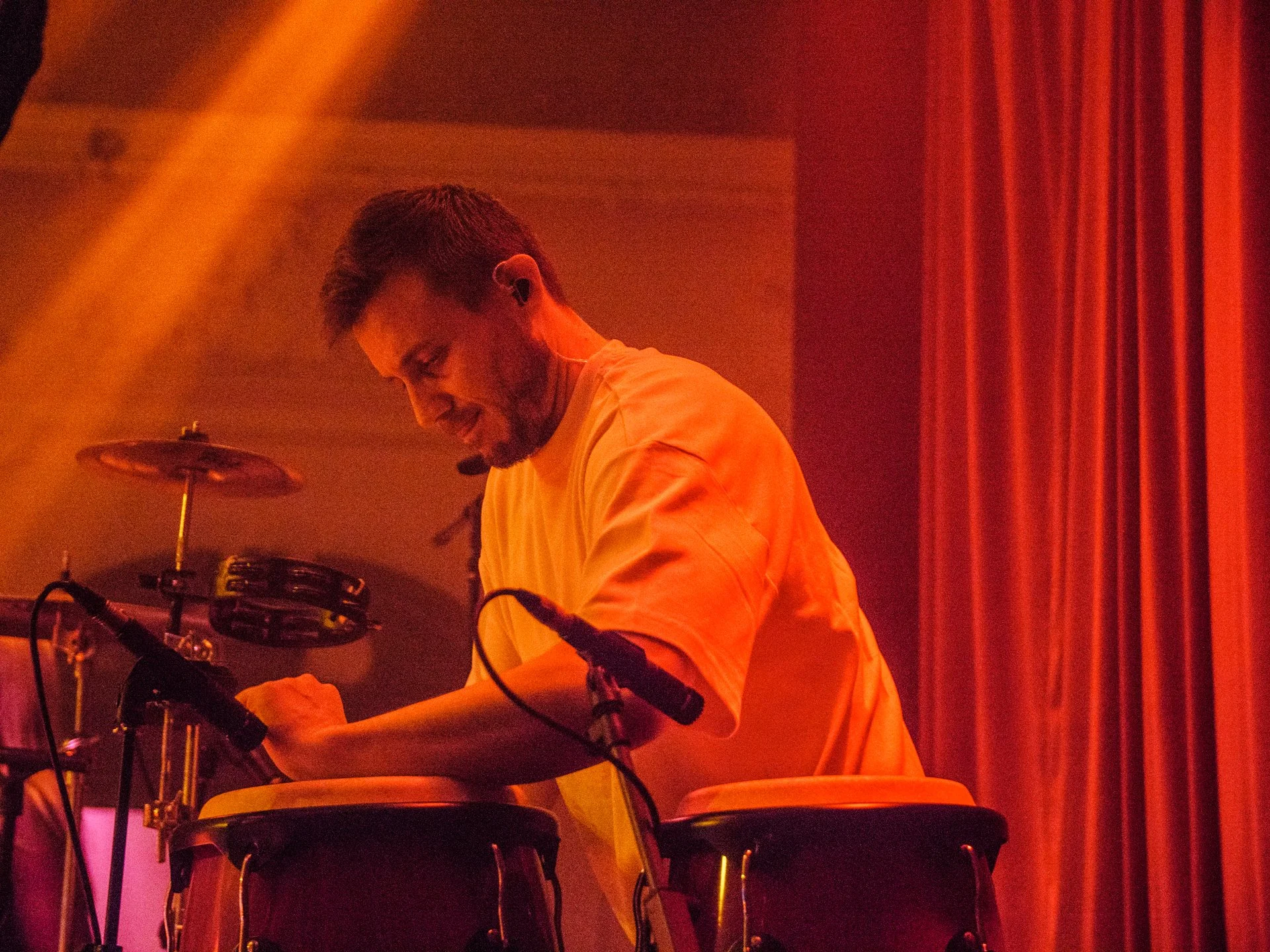 A man playing conga drums on stage, smiling and wearing a yellow shirt, illuminated with orange and red stage lighting.