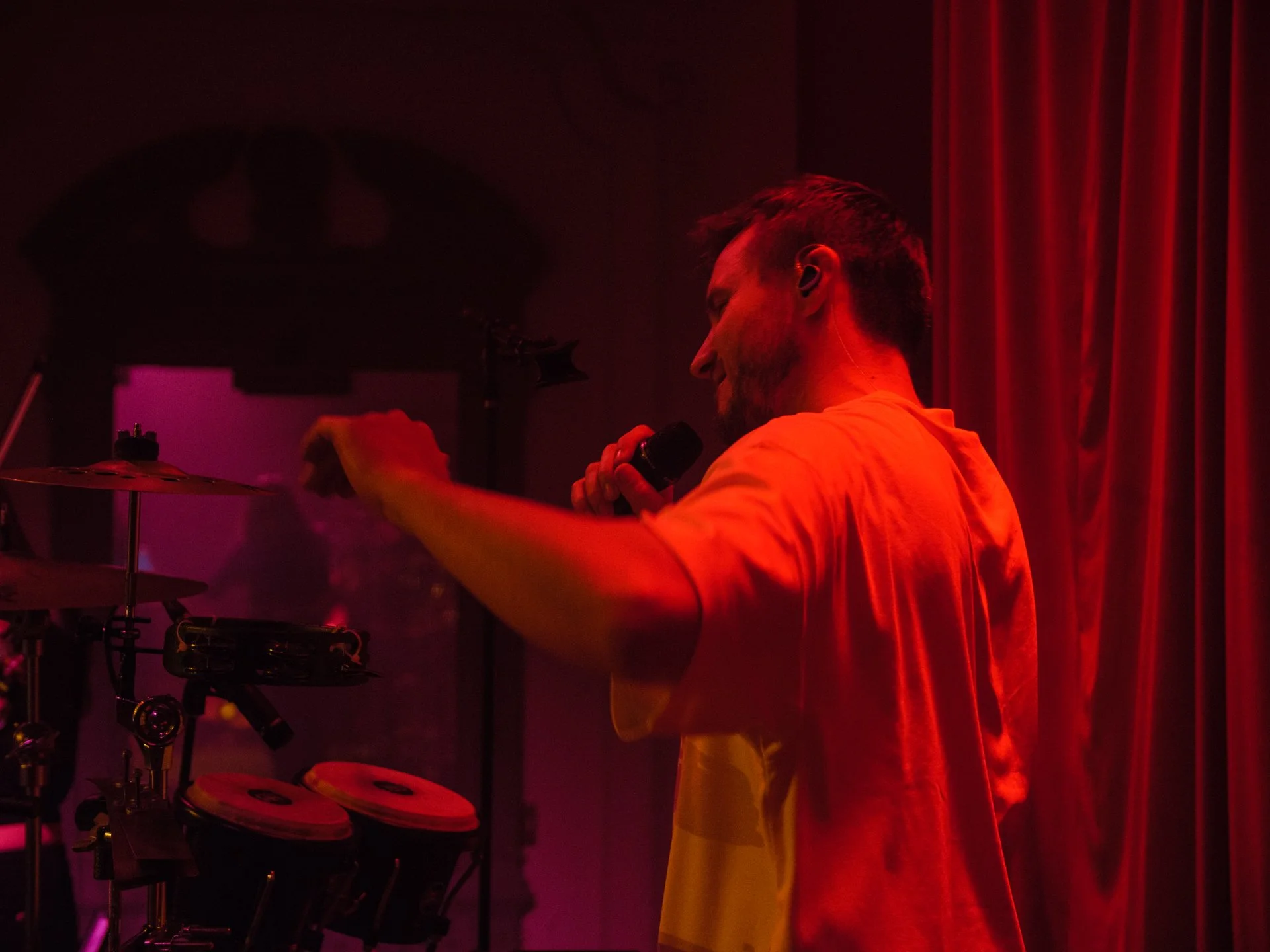 A male musician with a beard, holding a microphone in one hand, playing electronic drums on stage with red lighting and curtains in the background.