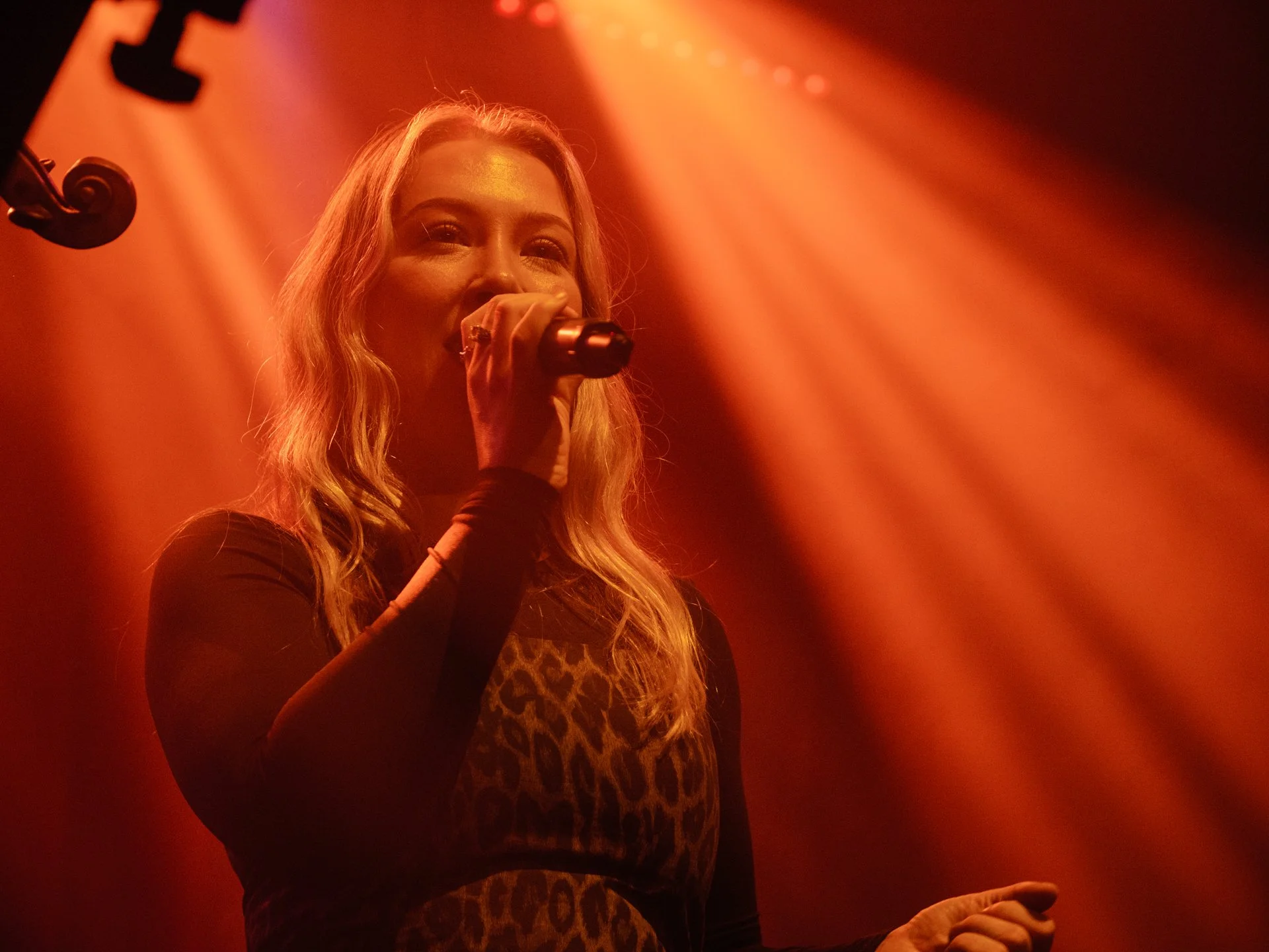 A woman with long blonde hair singing into a microphone on a stage with orange lighting.