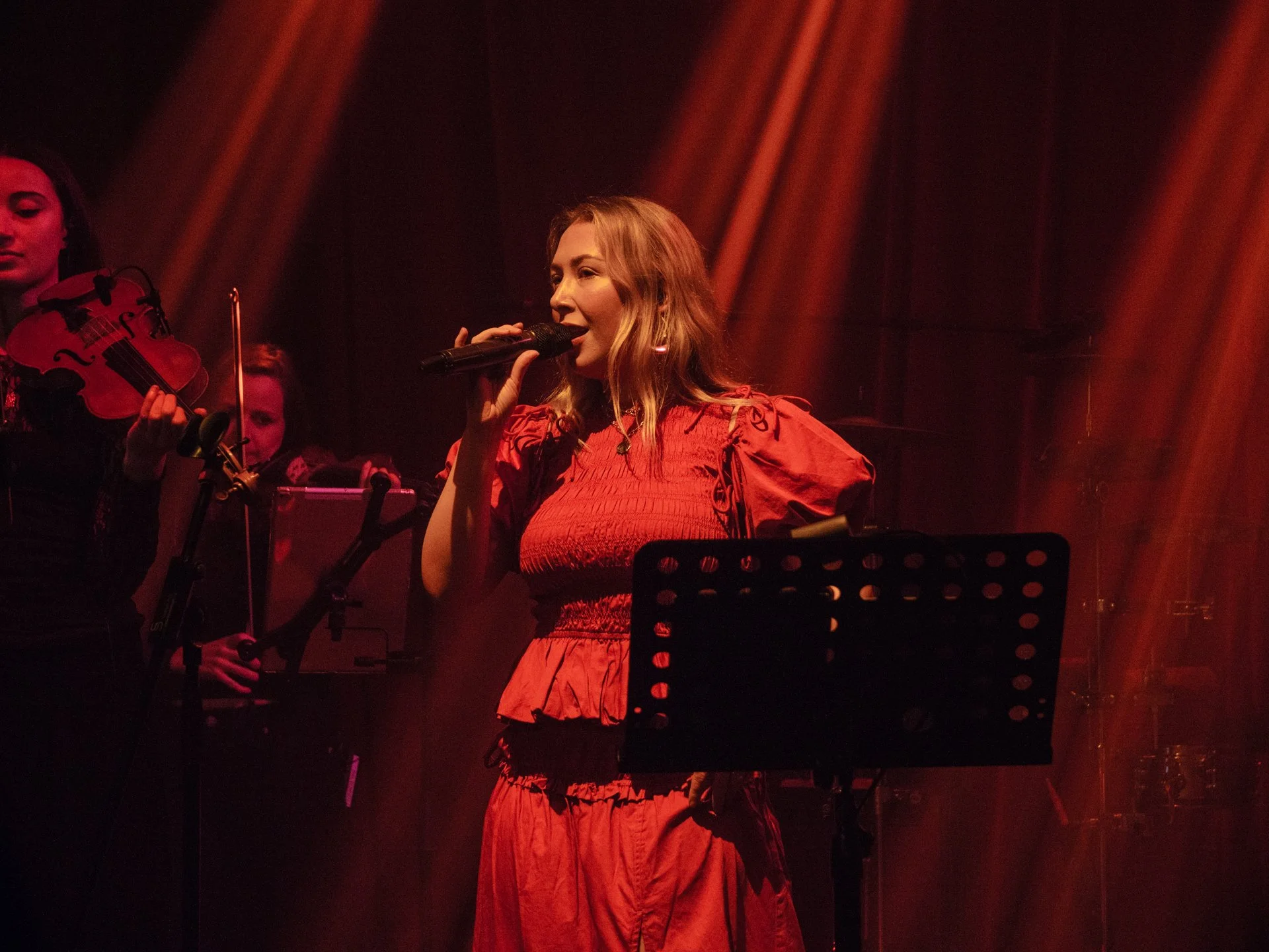 A woman singing into a microphone on stage, dressed in a red outfit, with violinists in the background, illuminated by warm stage lights.