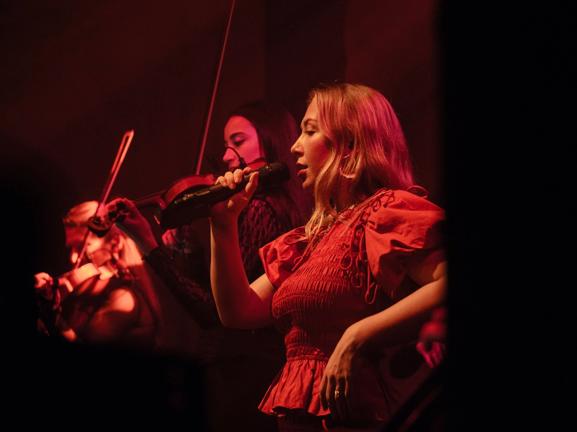 Two women performing on stage, one playing a violin and the other singing into a microphone, with red stage lighting.