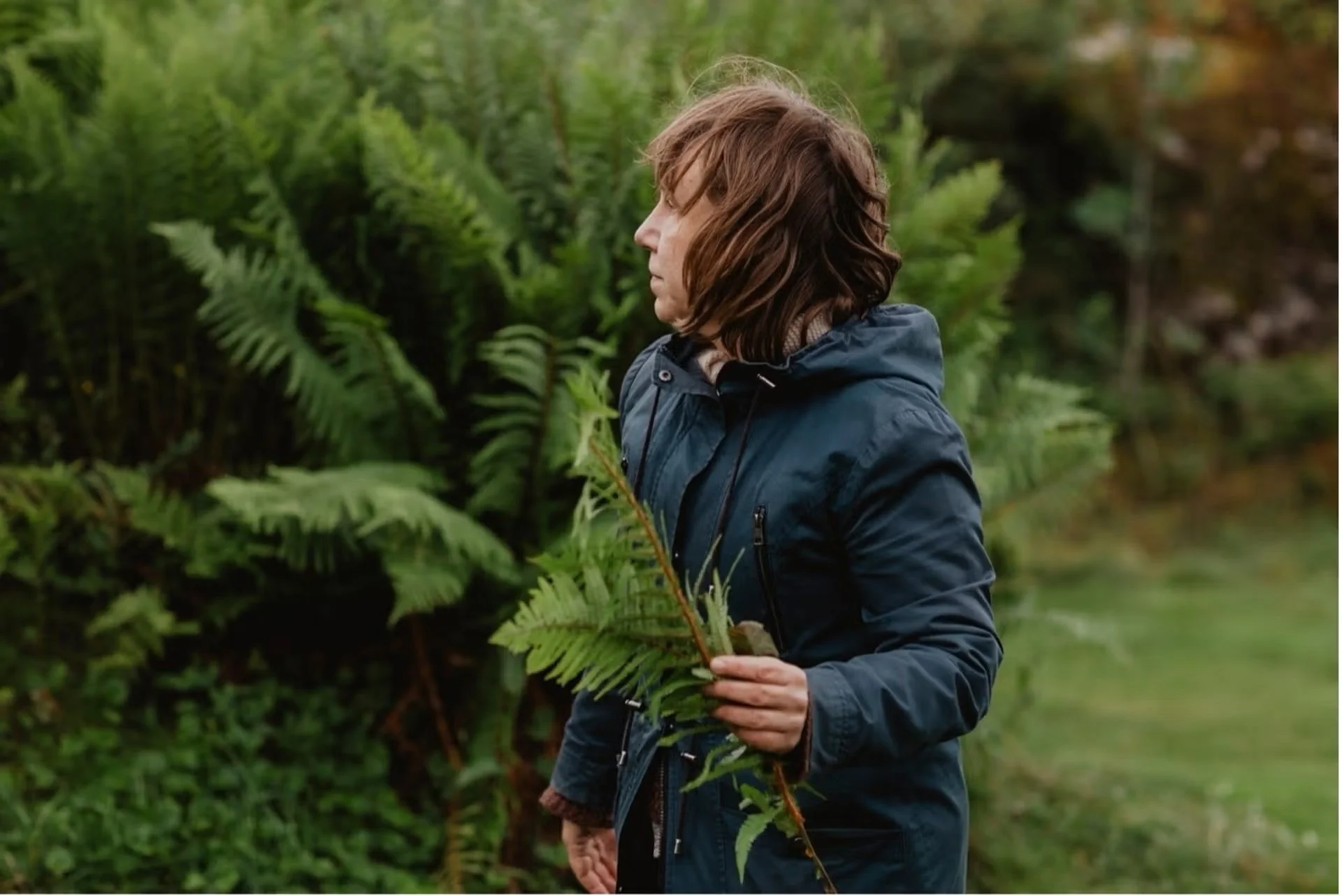 A woman with shoulder-length brown hair wearing a dark blue jacket holding a fern leaf in a lush, green outdoor setting with ferns and trees.