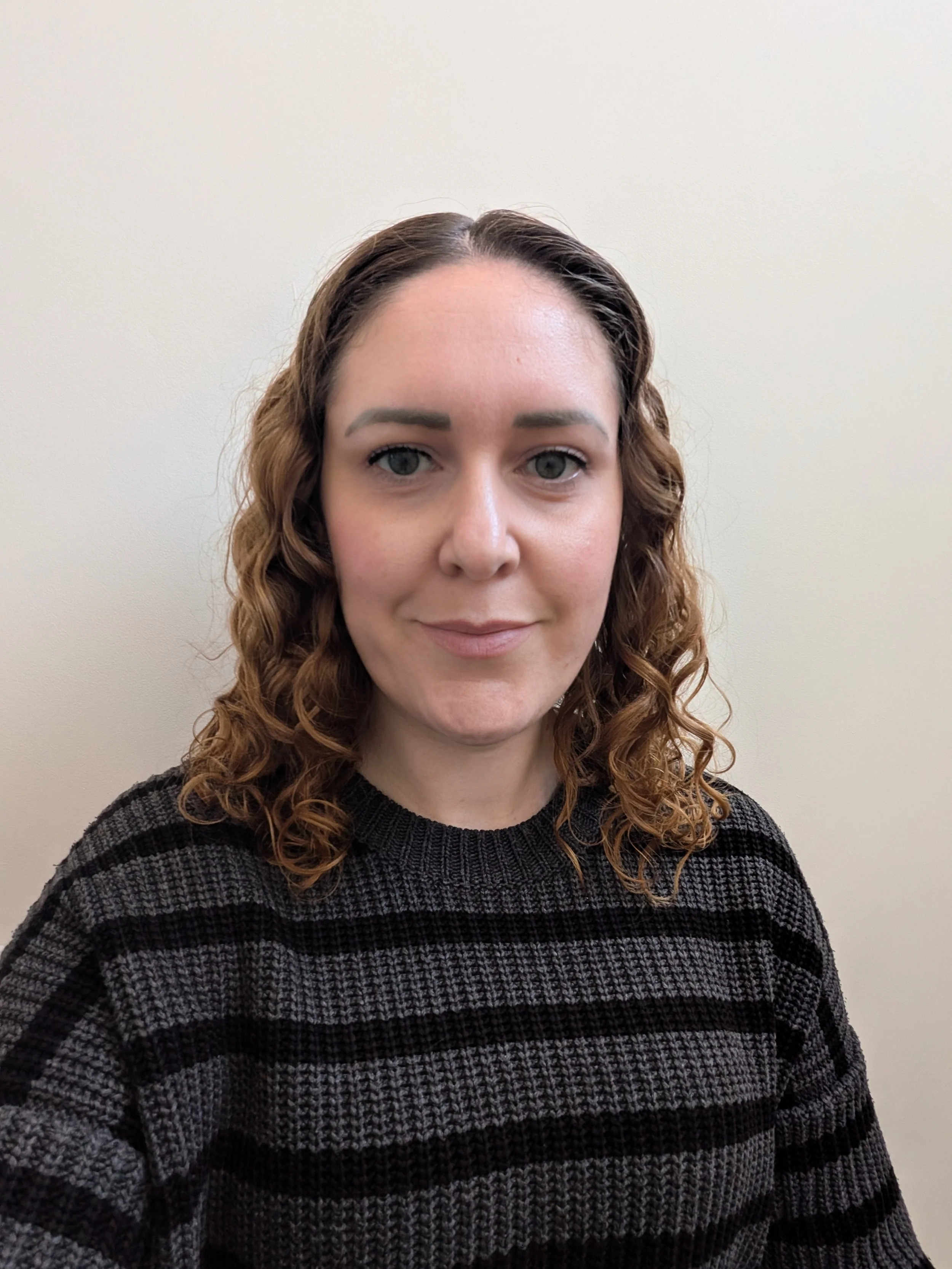 A woman with curly brown hair, wearing a black and gray striped sweater, standing against a plain beige wall.
