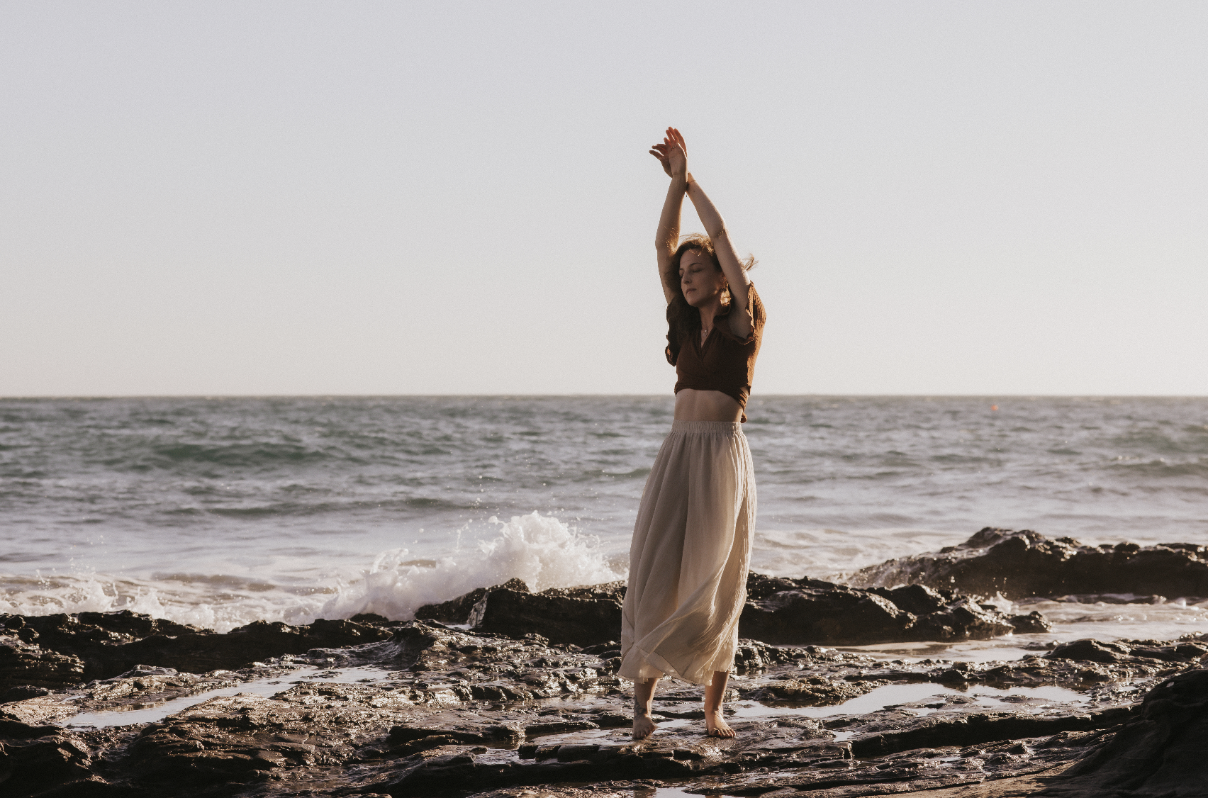 Frau tanzt barfuß auf Felsen am Strand bei Sonnenuntergang.