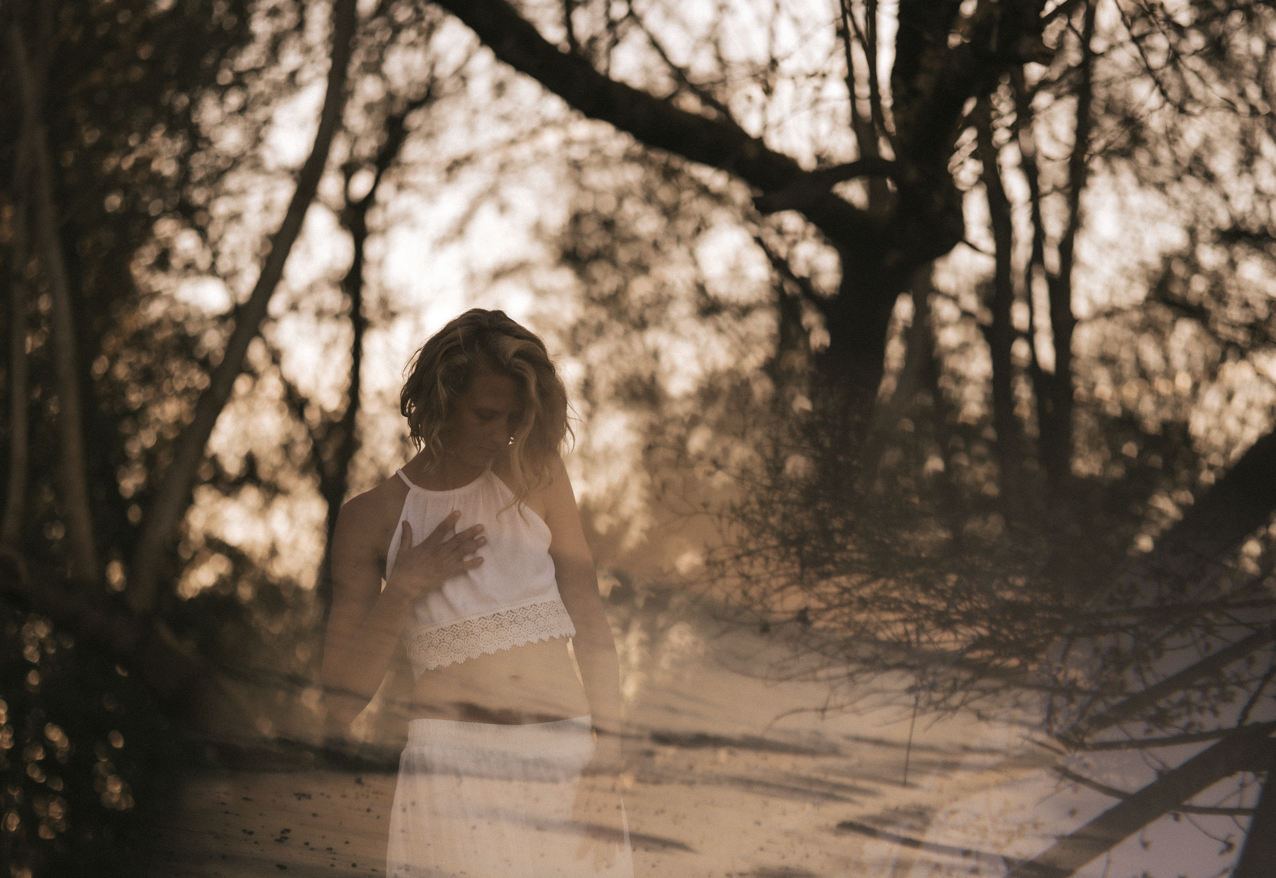 Eine Frau steht in einem Wald bei Sonnenuntergang, sie trägt ein weißes Crop-Top und einen weißen Rock, die Hand liegt auf ihrer Brust, während sie nach unten schaut.