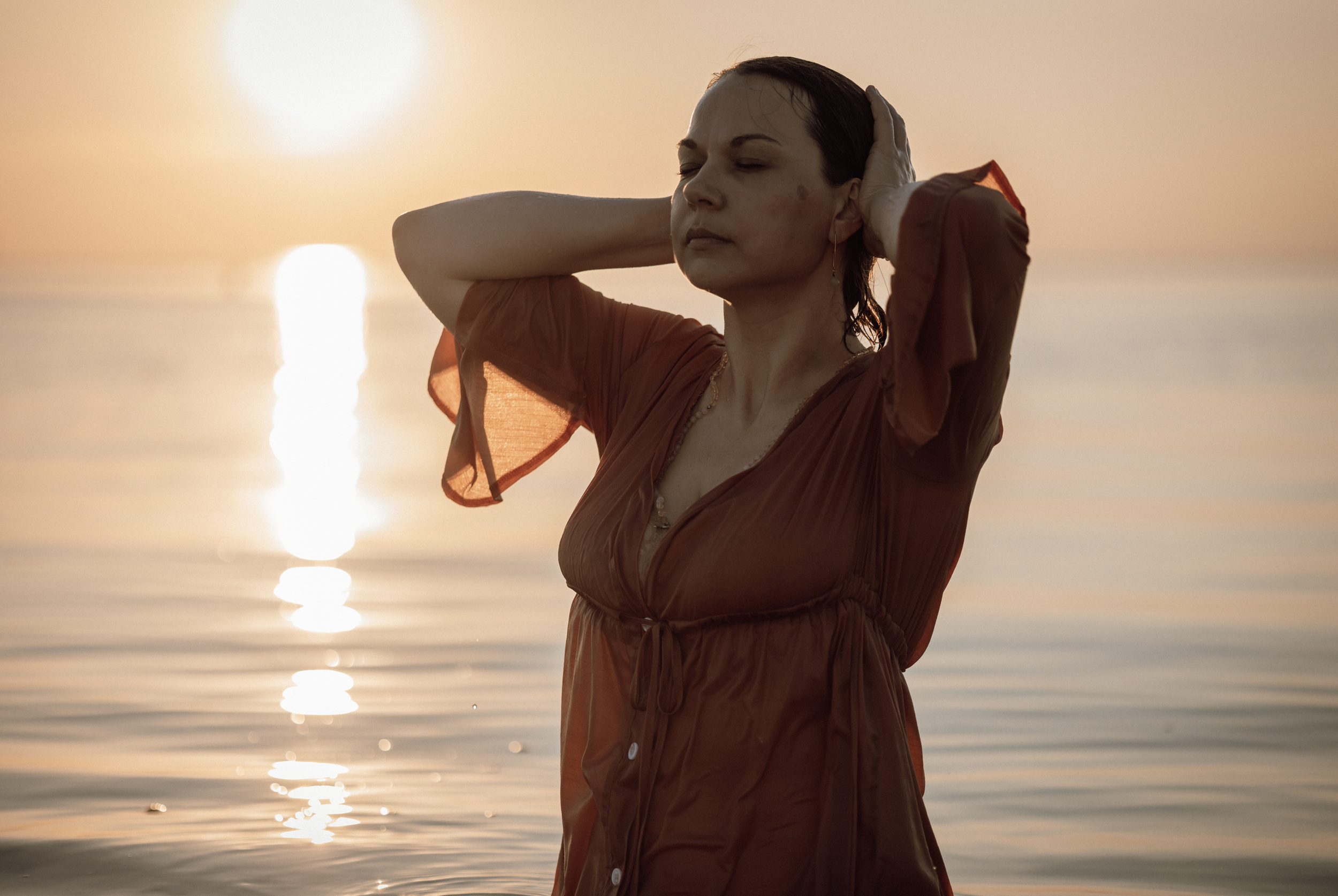 Frau im roten Kleid steht am Wasser bei Sonnenuntergang, Hände im Haar, mit Blick nach oben, ruhige Stimmung.