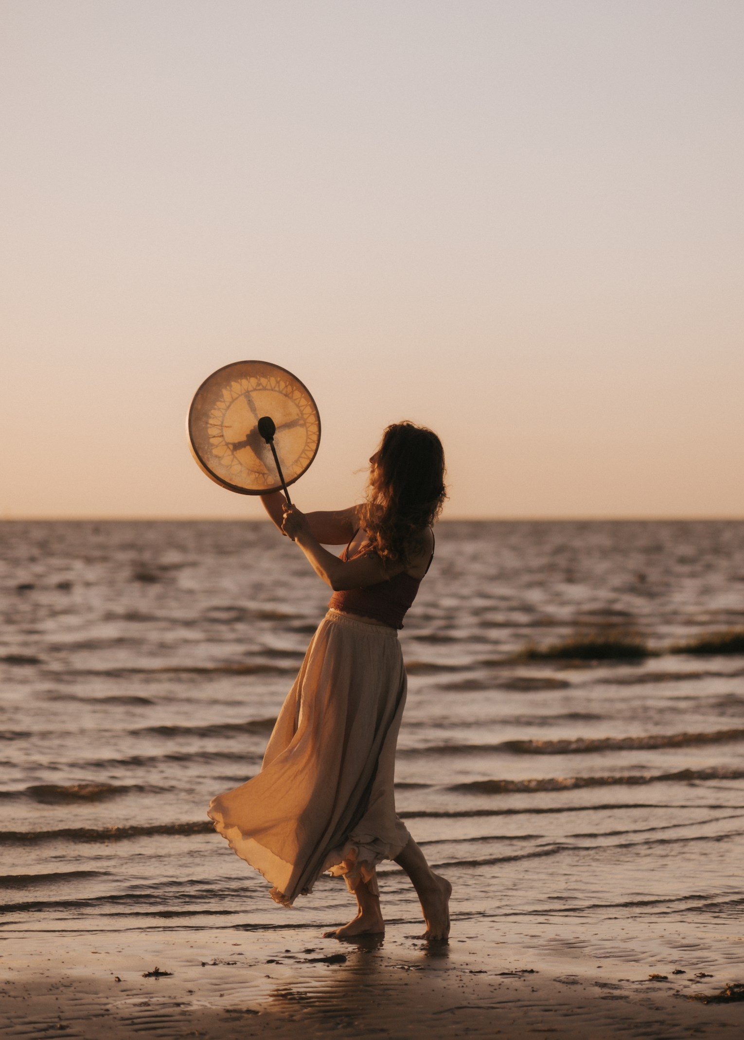 Frau tanzt am Strand bei Sonnenuntergang, hält ein Gonginstrument in der Hand.