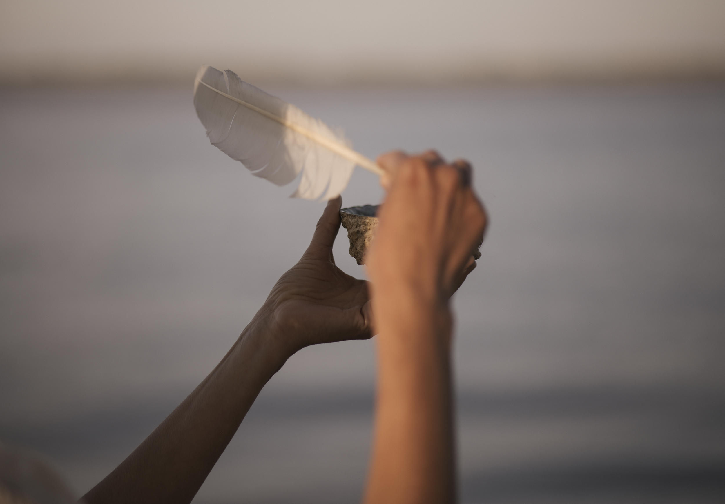 Nahaufnahme von einer Hand, die einen weißen Federkiel auf einem Felsen hält, mit einem ruhigen Wasser im Hintergrund bei Sonnenuntergang.