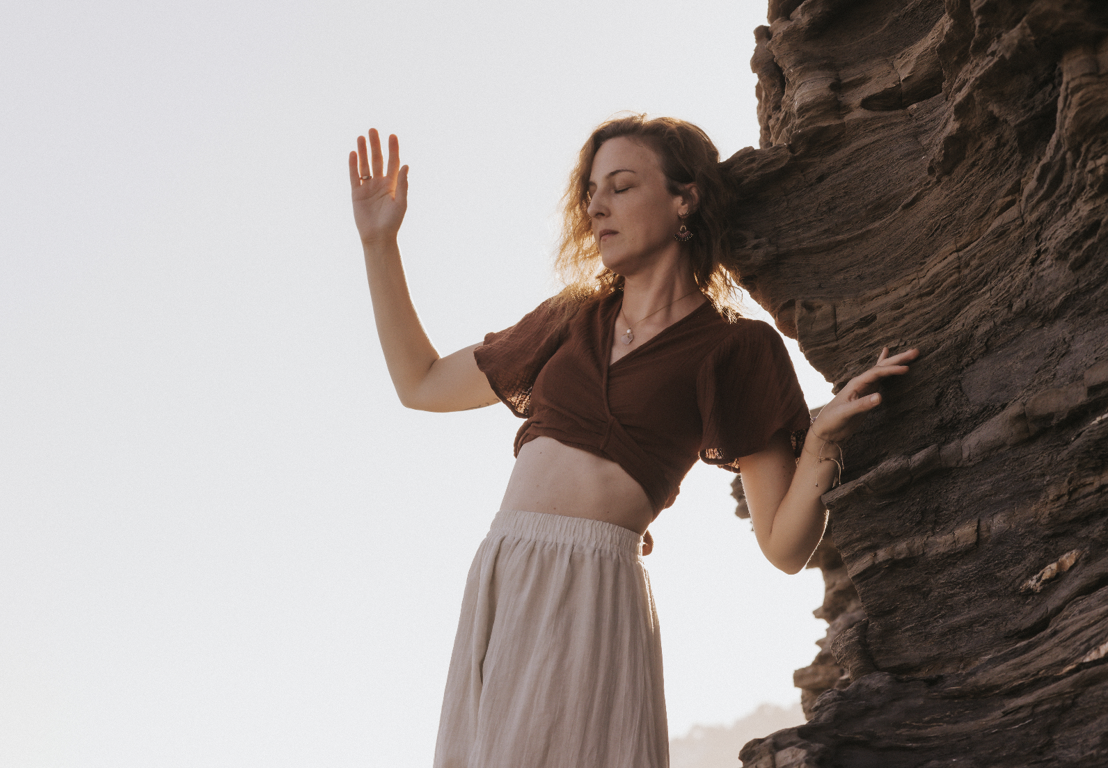 Eine junge Frau steht neben einer Felswand mit den Augen geschlossen, während sie die Hand in die Luft hebt und die andere Hand gegen den Felsen stützt. Sie trägt ein braunes Oberteil und eine beige Hose.