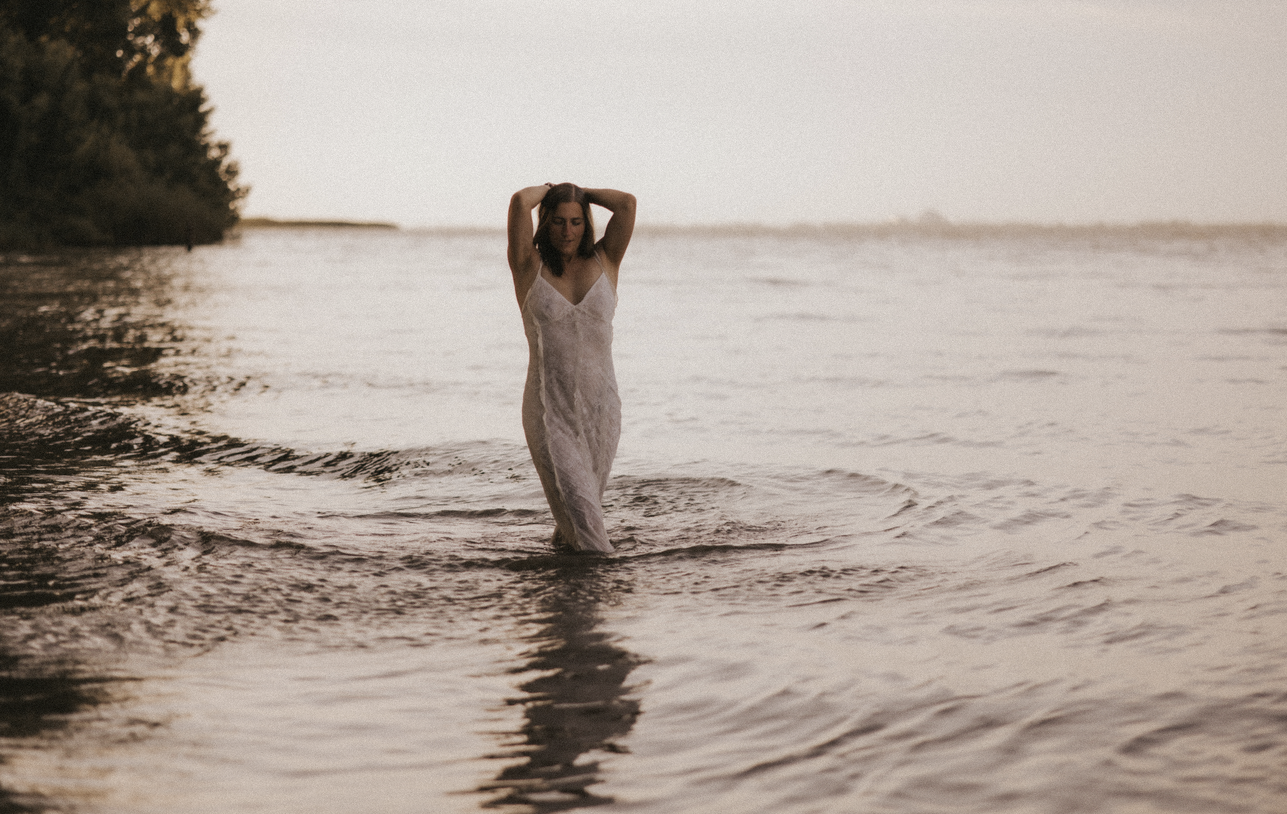 Junge Frau in weißem Kleid steht im Wasser am Strand während Sonnenuntergang.