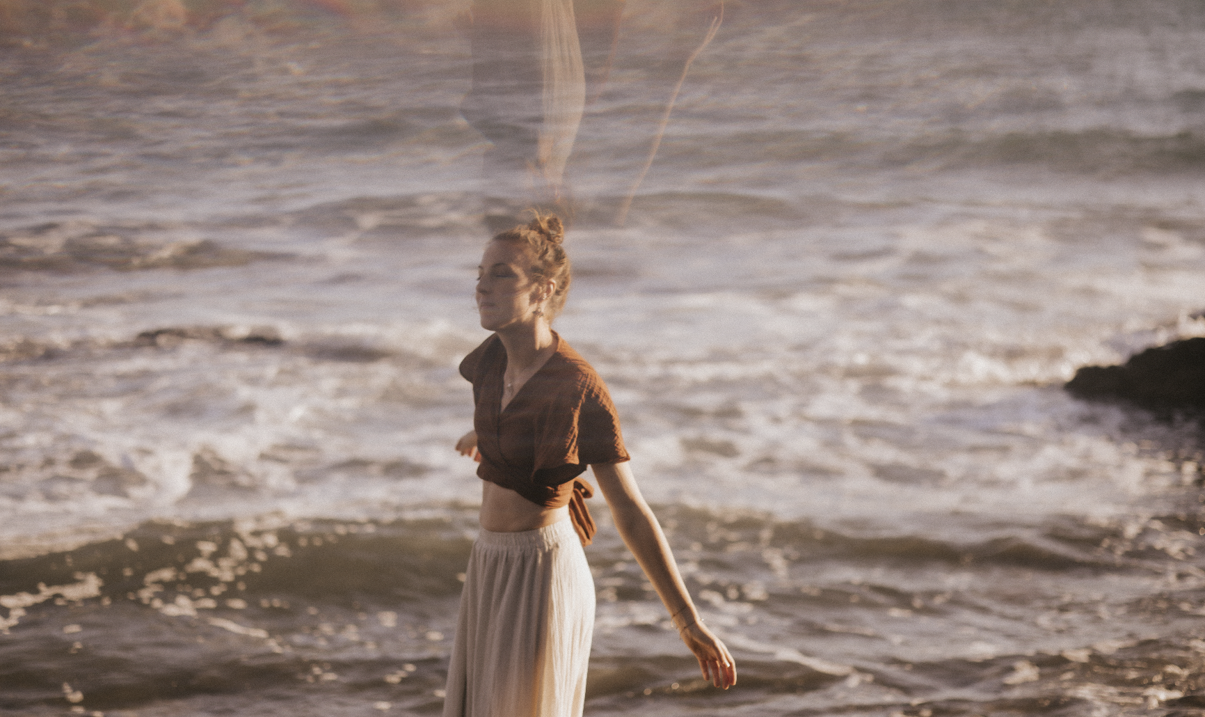 Eine Frau steht entspannt am Strand mit Blick auf das Meer, trägt einen braunen Knoten-Shirt und einen langen, hellen Rock bei Sonnenuntergang.
