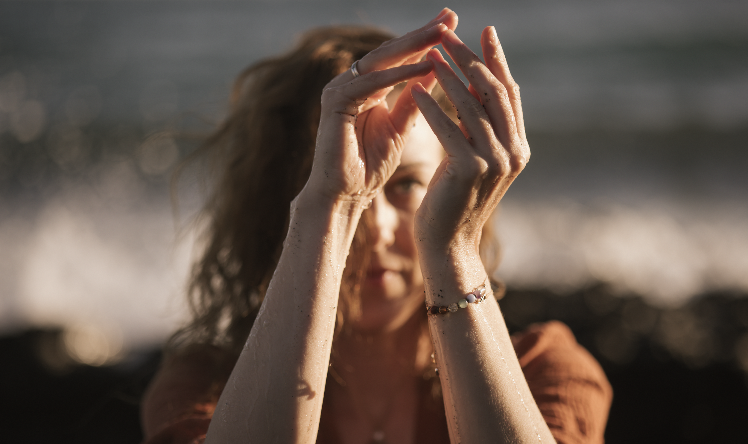 Frau mit nassen Haaren, die am Strand bei Sonnenuntergang mit den Händen ein imaginäres Dreieck formt.