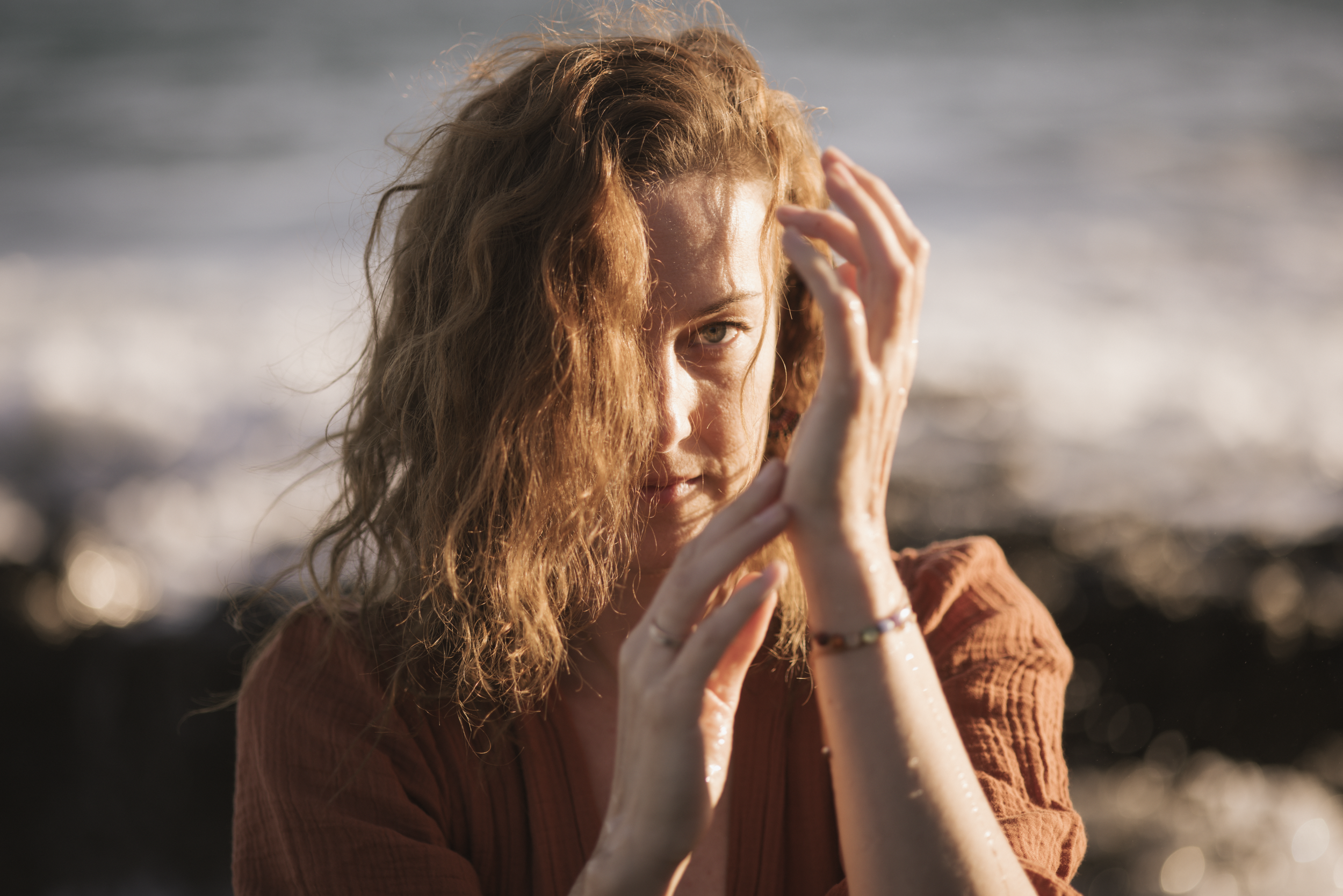 Eine junge Frau mit langen, lockigen roten Haaren, die bei Sonnenuntergang am Strand in das Meer schaut und die Hände an das Gesicht legt.