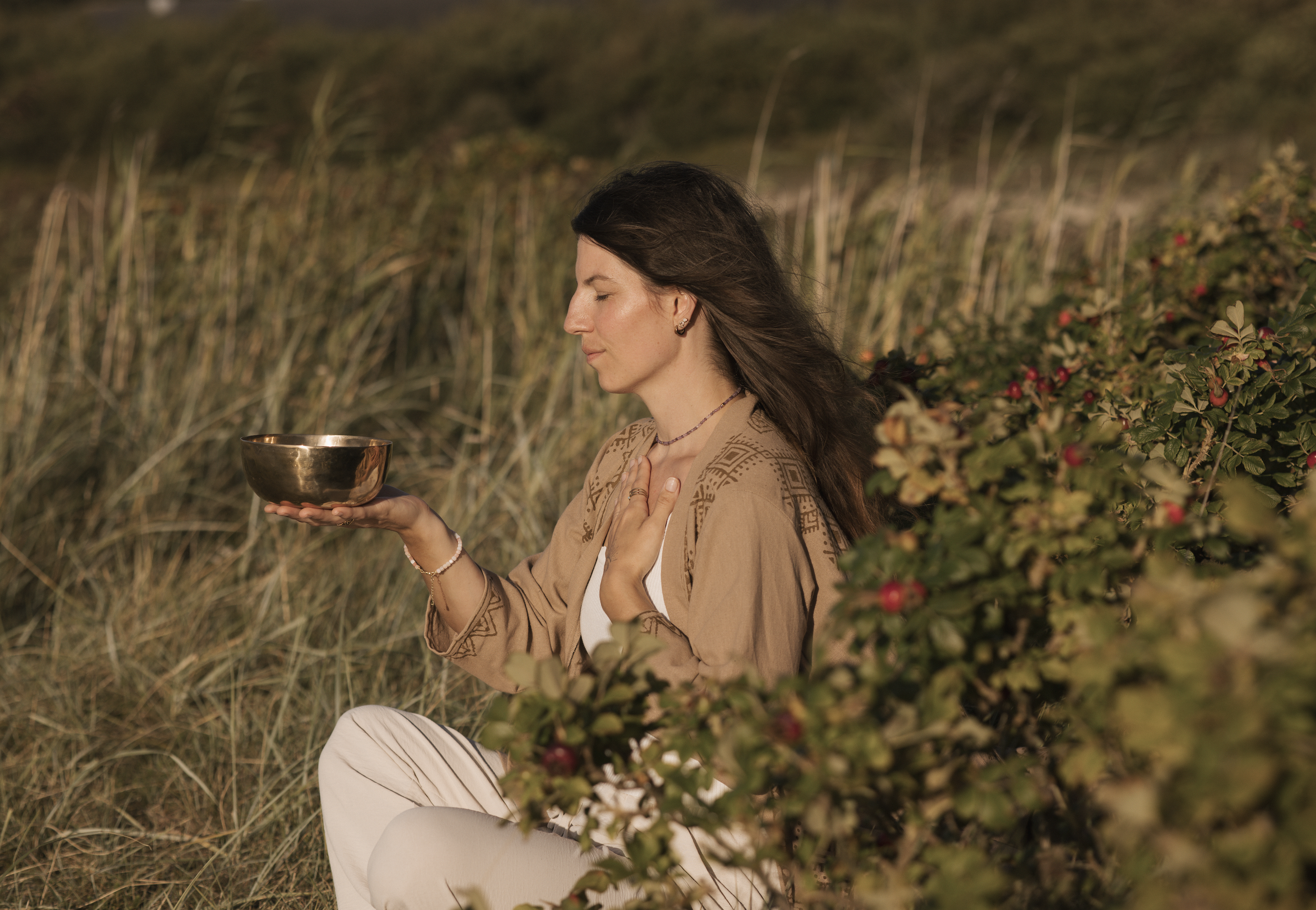 Eine Frau sitzt im Freien neben Buchsbaum und Blumen, hält eine Metall-Schale in der Hand und hat die Augen geschlossen, wirkt meditativ.