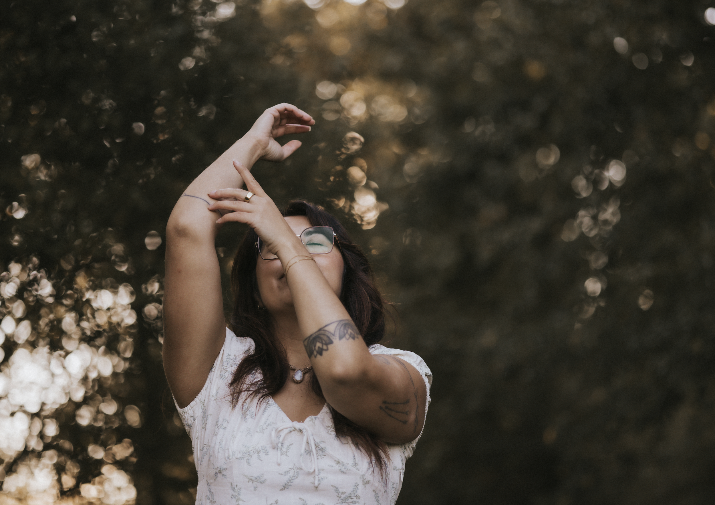 Junge Frau mit dunklen Haaren, Brille und Tattoos posiert mit verschränkten Armen vor einer unscharfen Baum- und Sonnenlicht-Hintergrund bei Sonnenuntergang