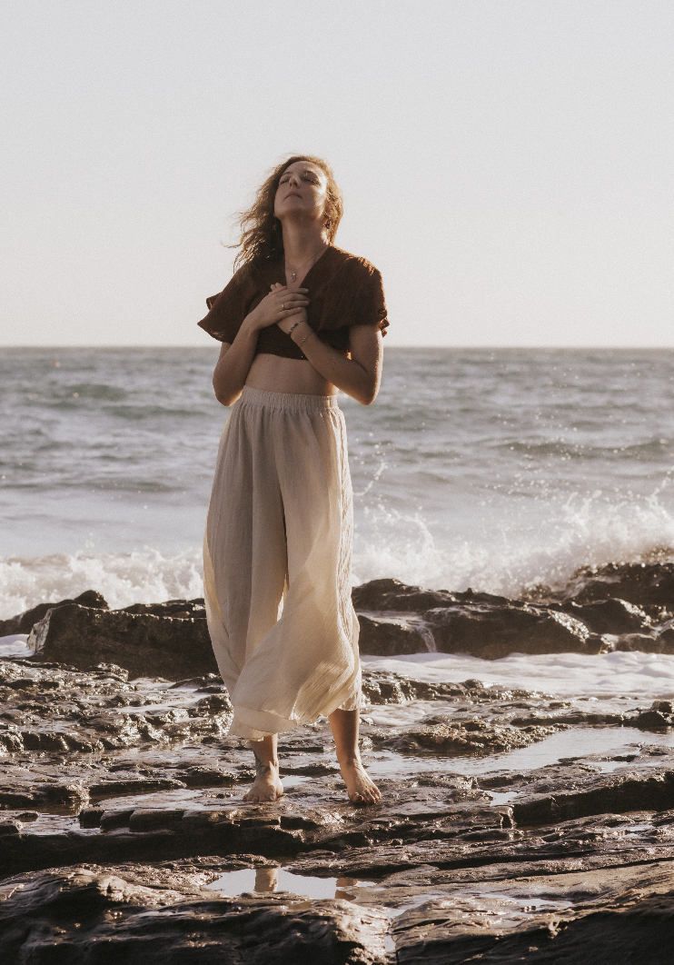 Frau steht barfuß auf einem felsigen Strand bei Sonnenuntergang, trägt weite beige Hosen und ein braunes Oberteil, mit Blick nach oben.