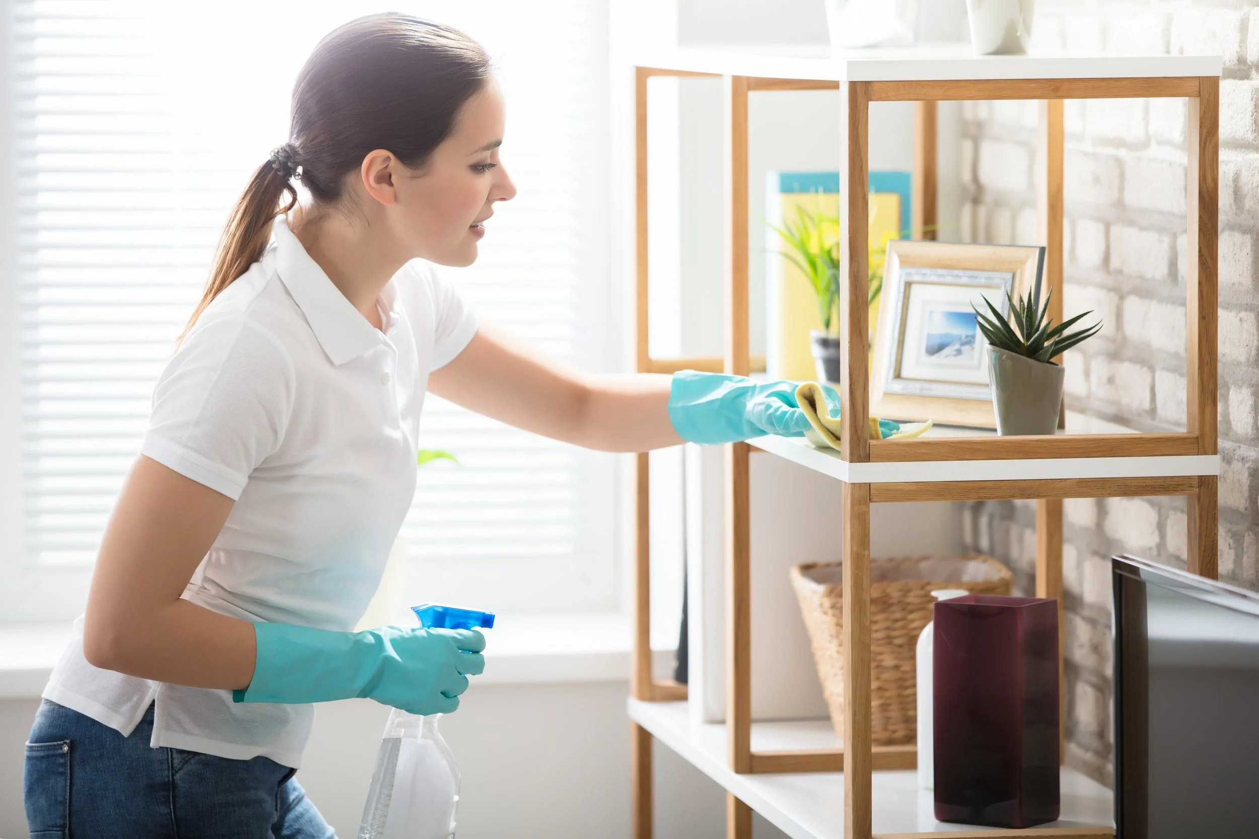 Woman cleaning a shelf with gloves and a spray bottle in a bright room.