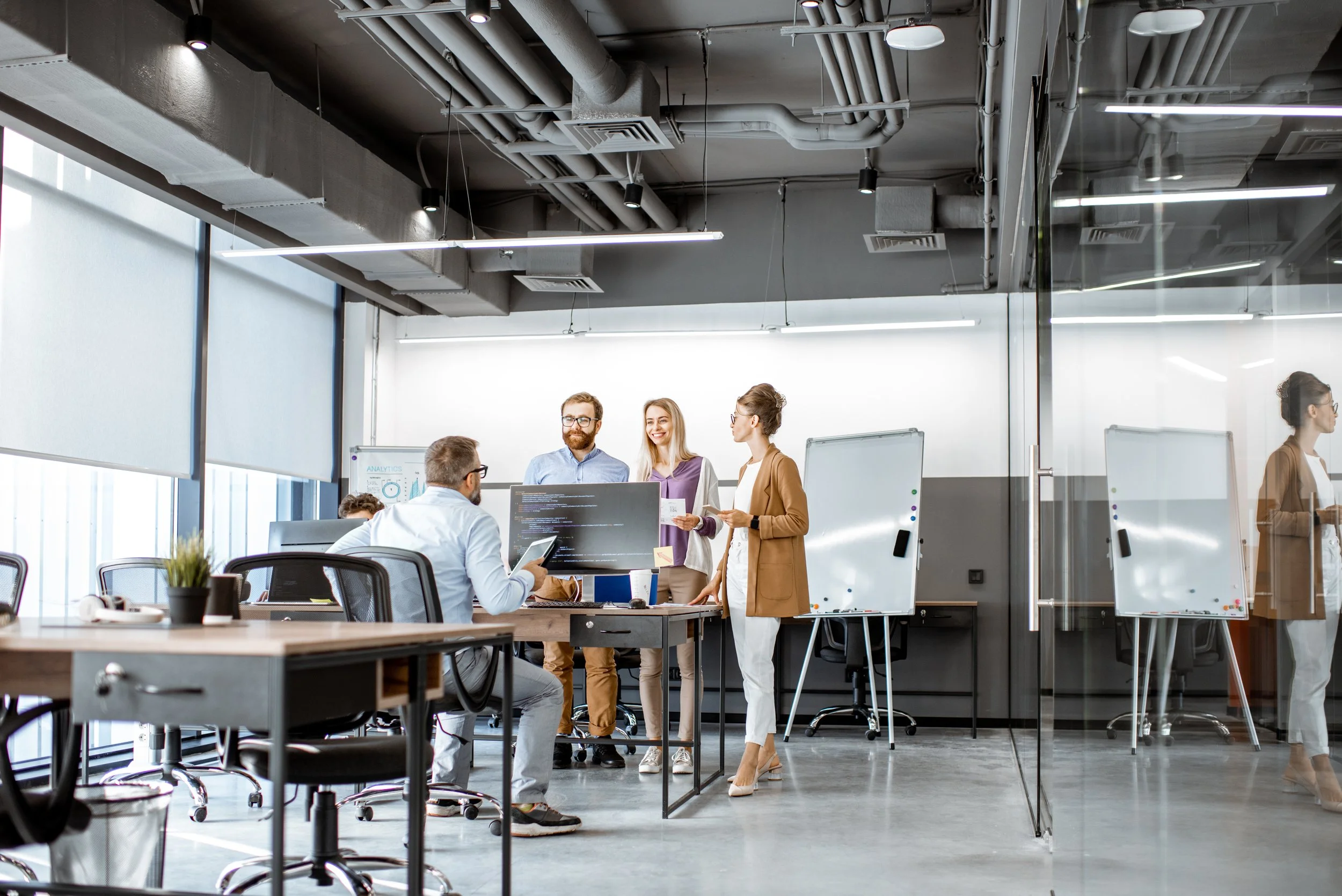 Business professionals in a modern office meeting room, standing and sitting around a table with a computer, whiteboards, and laptops, engaged in discussion.