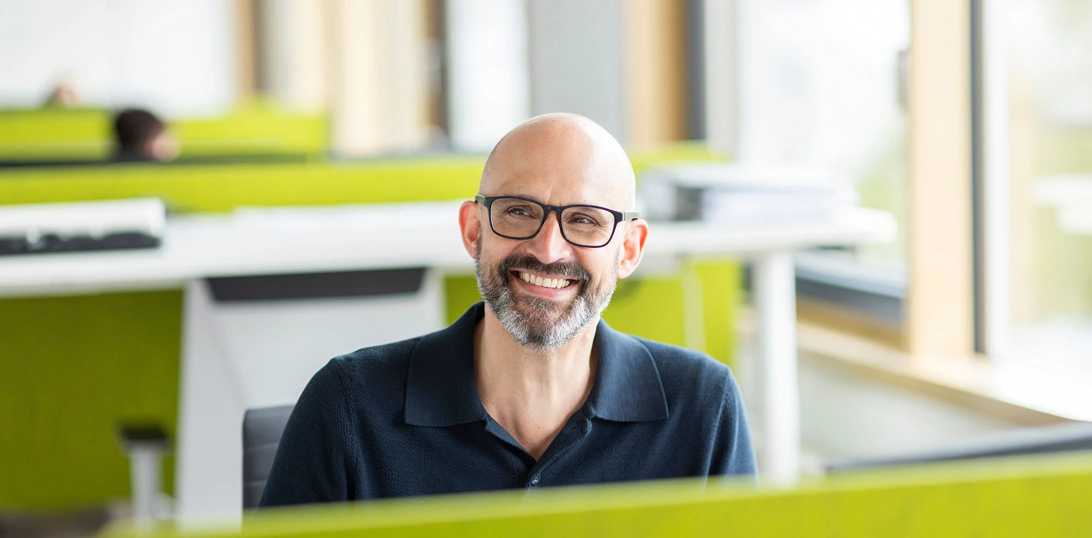 Ein lächelnder, älterer Mann mit Brille und Bart sitzt in einem modernen Büro mit grünen Akzenten und großen Fenstern im Hintergrund.
