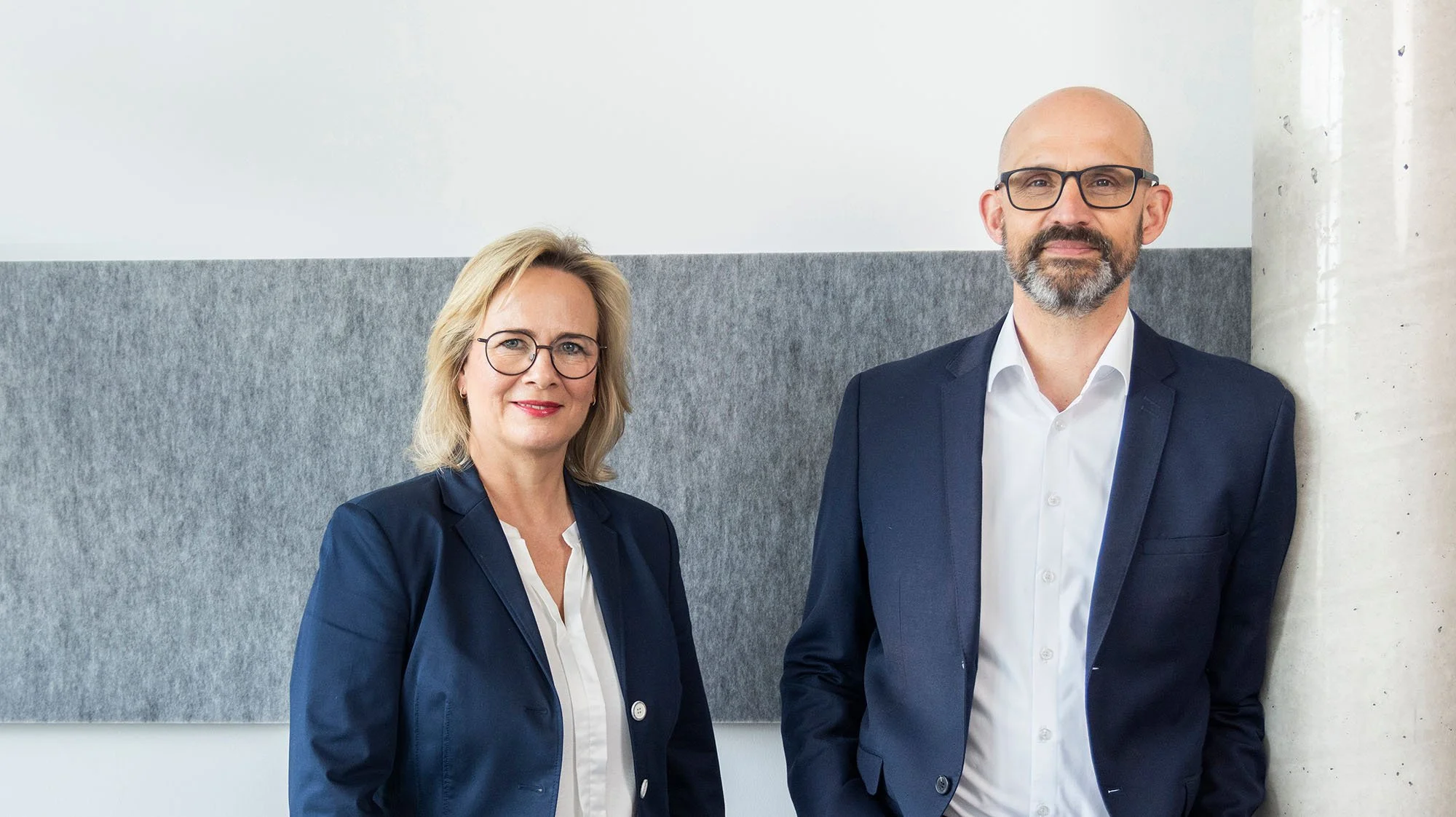 Isabel Brinkmann und Achim Bassa von kzp stehen nebeneinander vor einer grauen und weißen Wand, tragen formelle Kleidung und schauen in die Kamera.