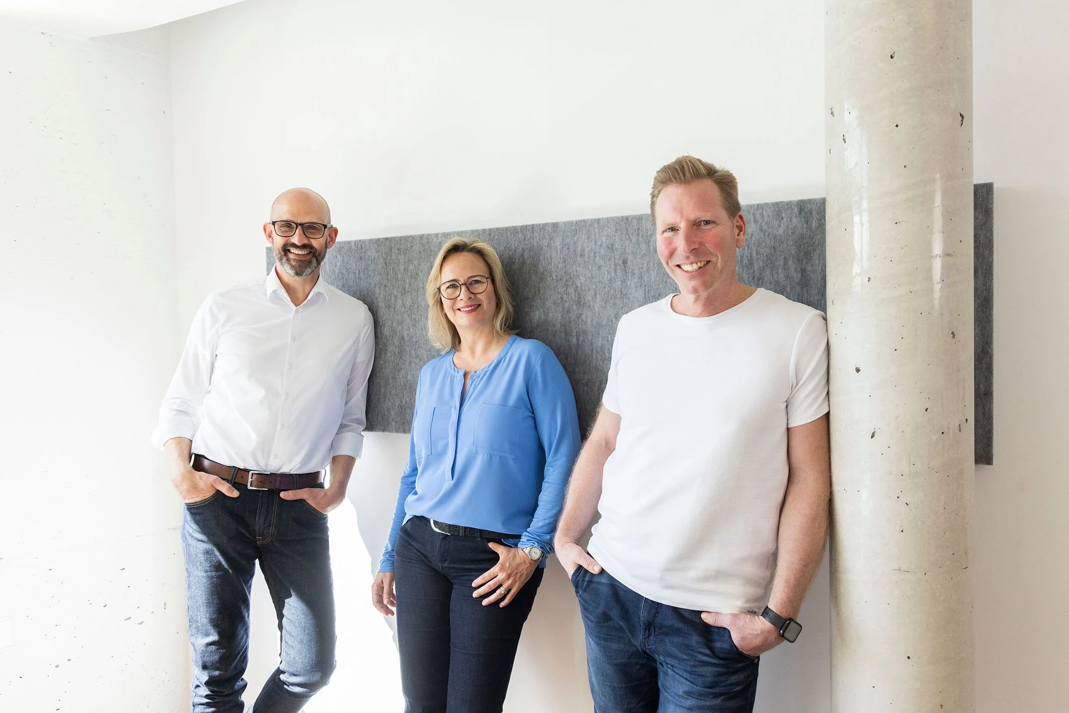 Drei Menschen stehen und lächeln vor einer weißen Wand, einer mit Brille, die anderen zwei ohne. Sie tragen legere Kleidung, der Mann rechts trägt eine weiße T-Shirt, die Frau in der Mitte eine blaue Bluse, ein Mann links ein weißes Hemd. Es ist Tageslicht im Raum.