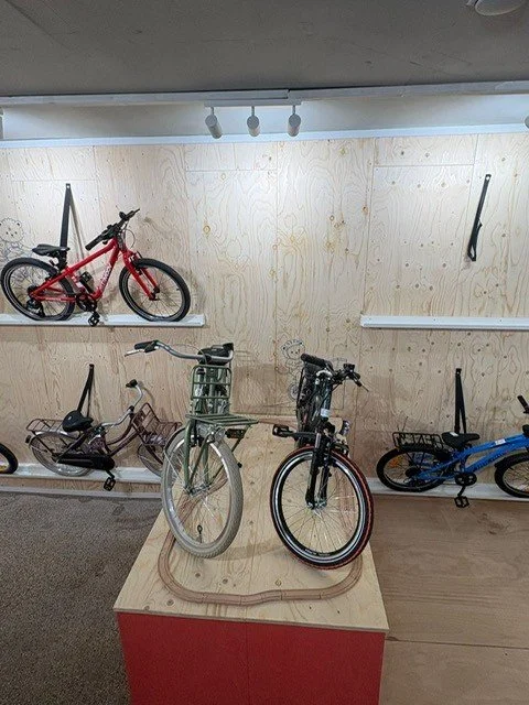 Three bicycles are displayed, two on the floor and one mounted on a wall in a room with plywood walls.