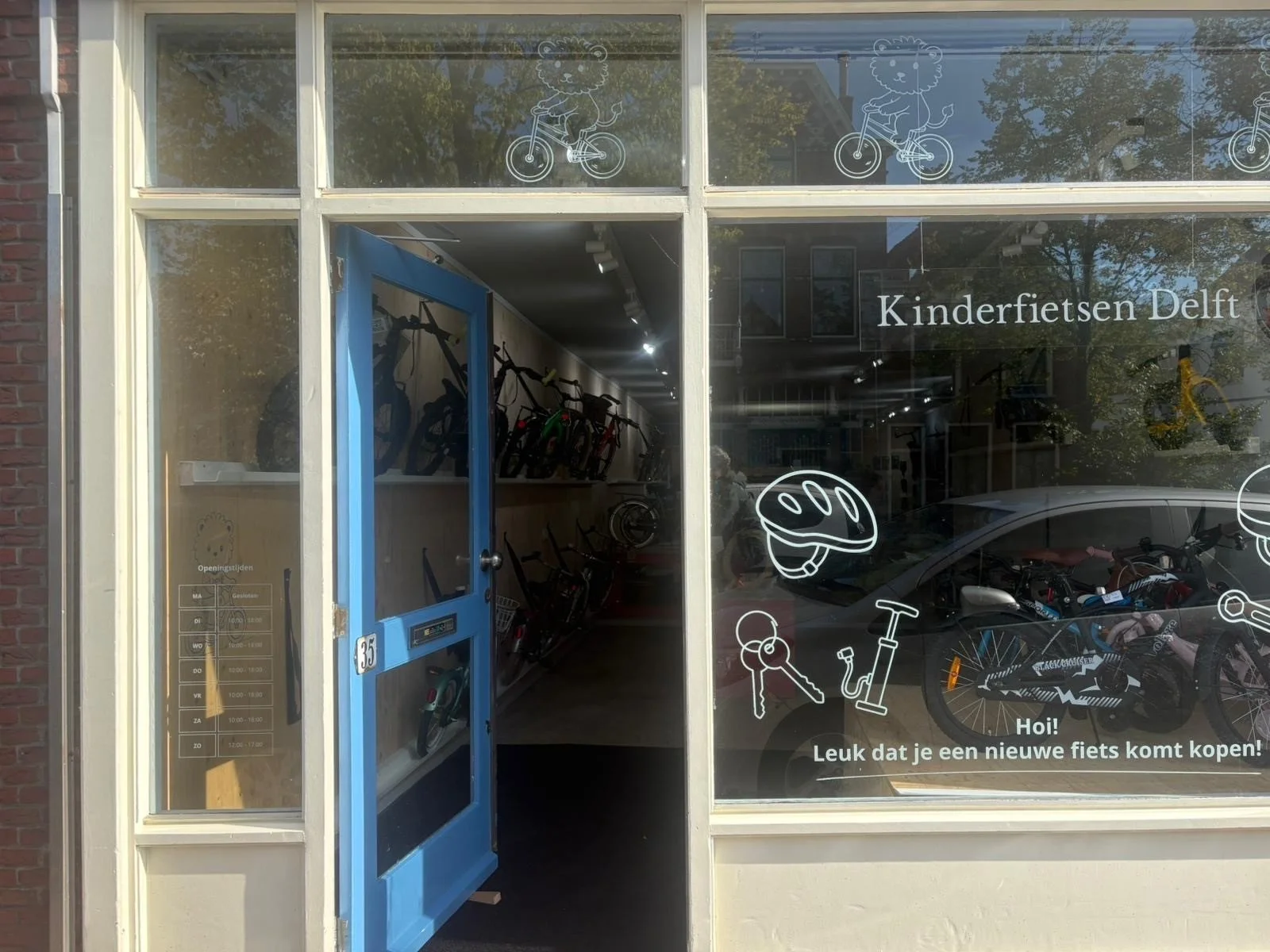 A storefront with glass windows and a blue door, showing a bicycle shop called 'Kinderfietsen Delft'. The window features line art of a lion riding a bicycle, a helmet, keys, and a scooter, along with the text 'Hoi! Leuk dat je een nieuwe fiets komt kopen!' which translates to 'Hi! Nice that you're coming to buy a new bike!'. Inside, several bikes are displayed on shelves and a bicycle is parked outside.