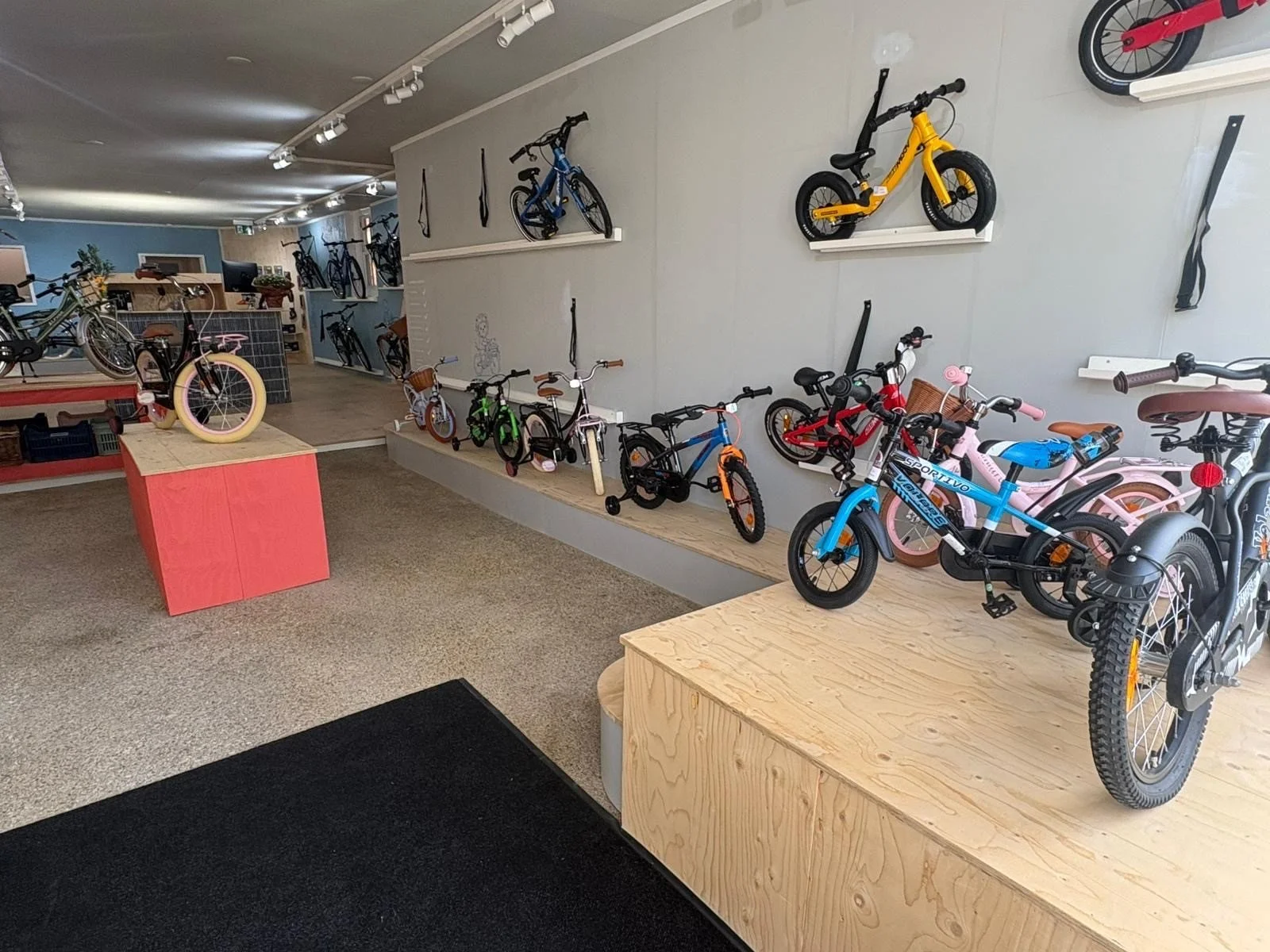 Indoor bike and scooter shop displaying various bicycles, balance bikes, and scooters on display shelves and platforms.