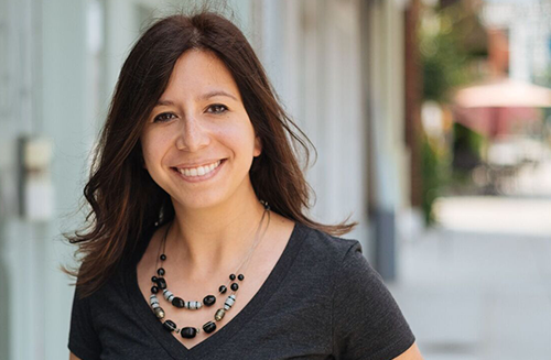 A smiling woman with shoulder-length dark hair wearing a black T-shirt and a layered black bead necklace standing outdoors on a sunny day.