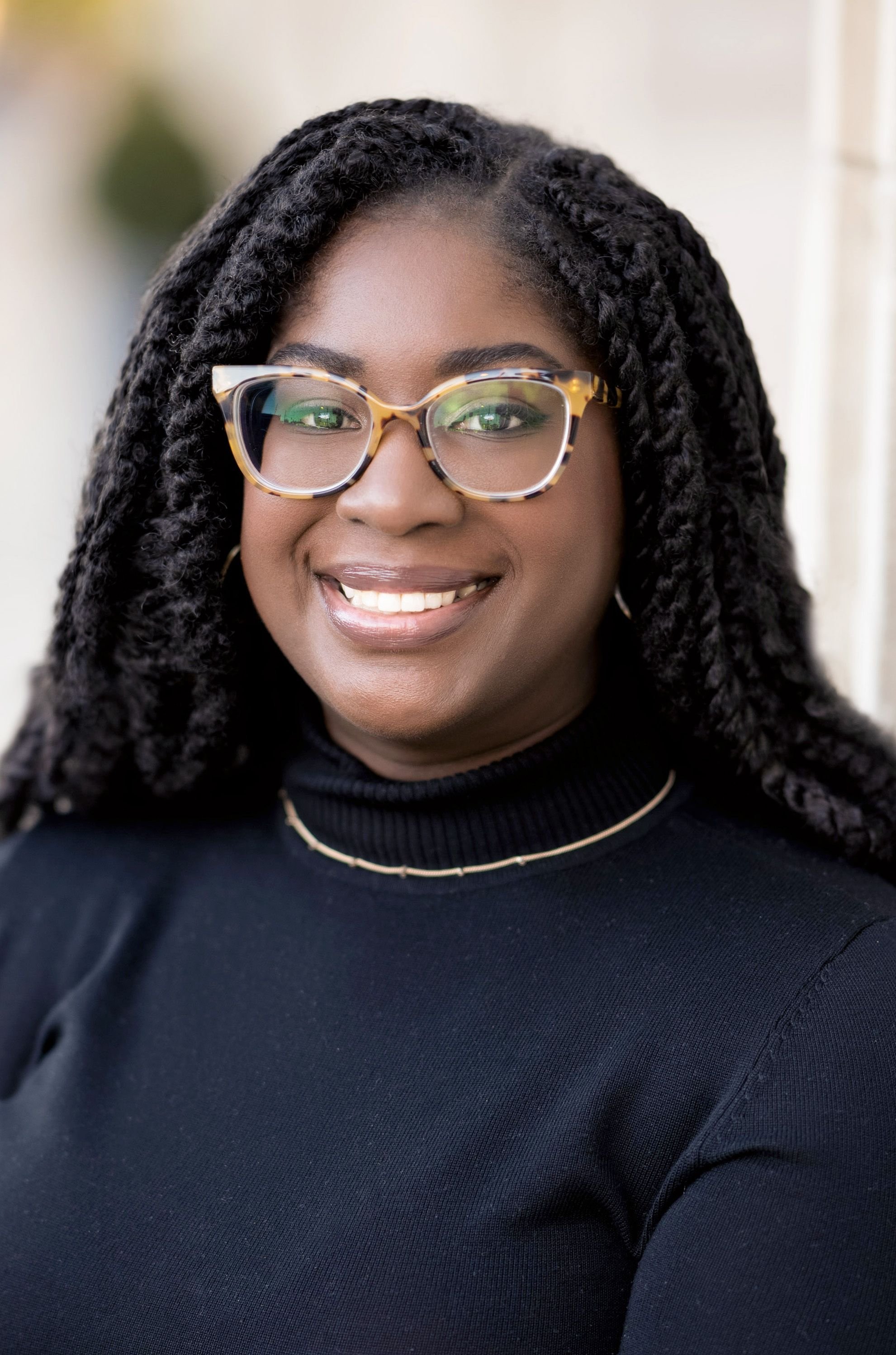 A woman with black curly hair wearing large tortoiseshell glasses, a black turtleneck top, a delicate gold necklace, and earrings, smiling at the camera.