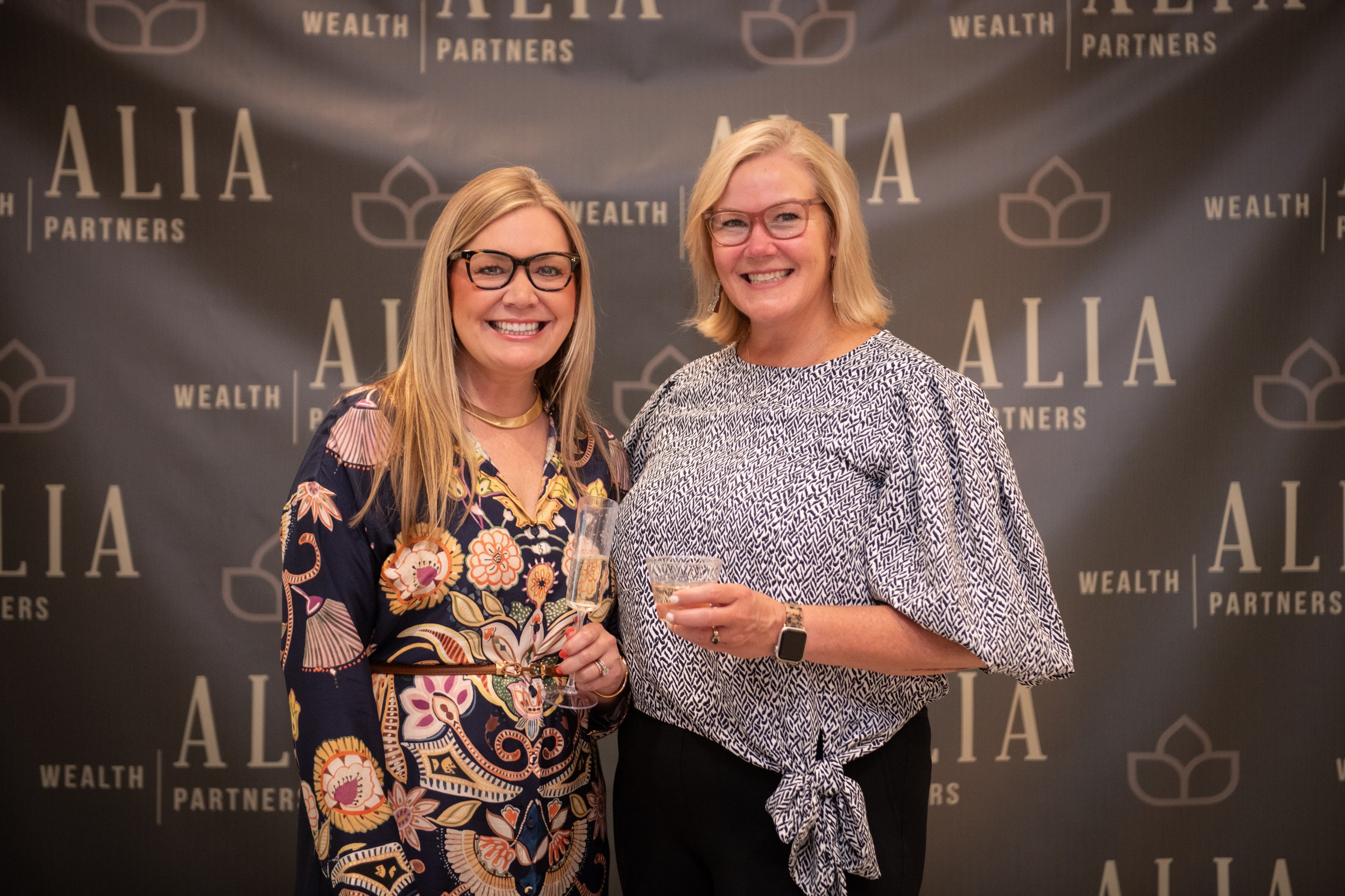 Two smiling women holding glasses of champagne at an event, standing in front of a backdrop with the words 'Alia Wealth Partners' written repeatedly.