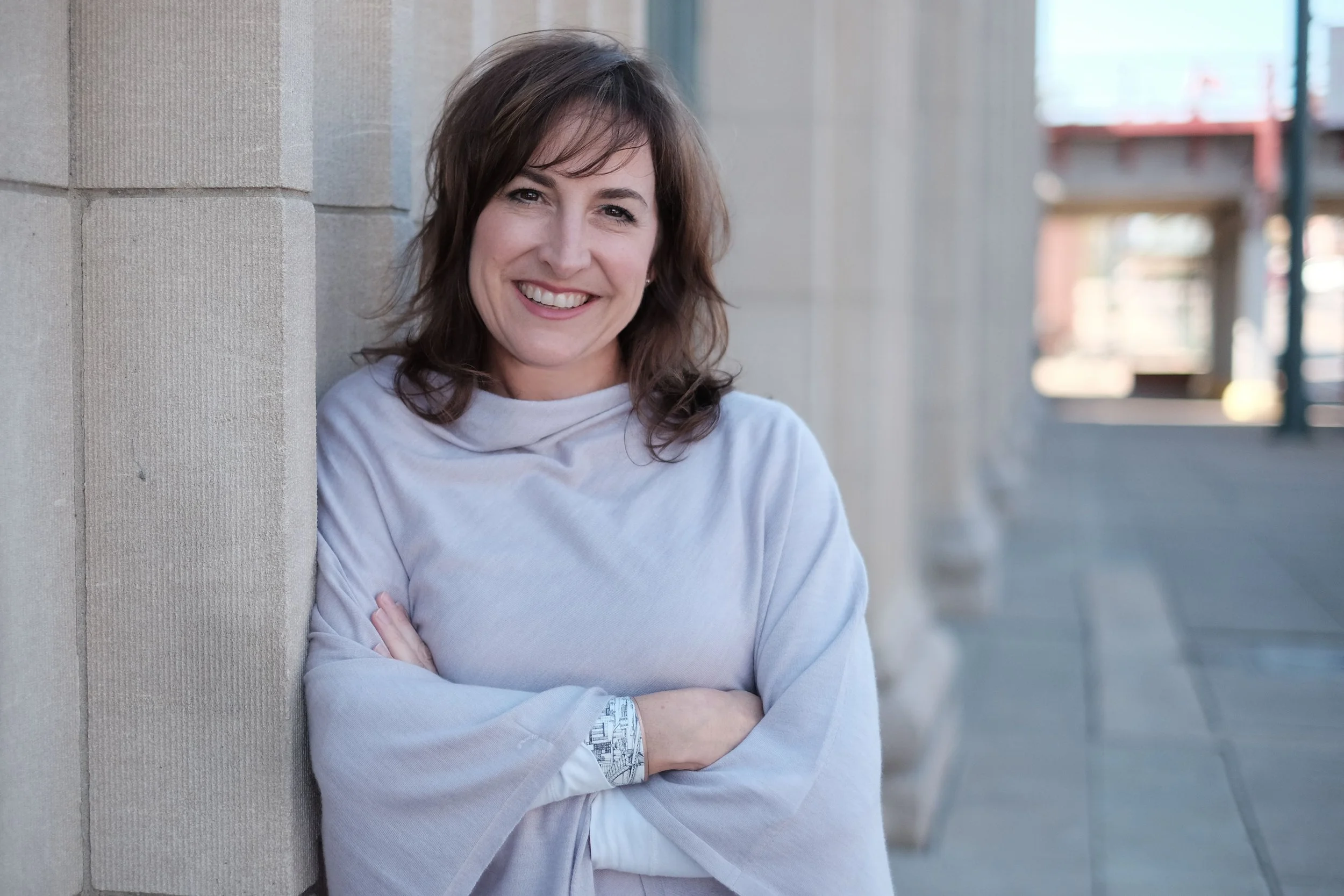 A woman with brown hair smiling, leaning against a brick wall outdoors.