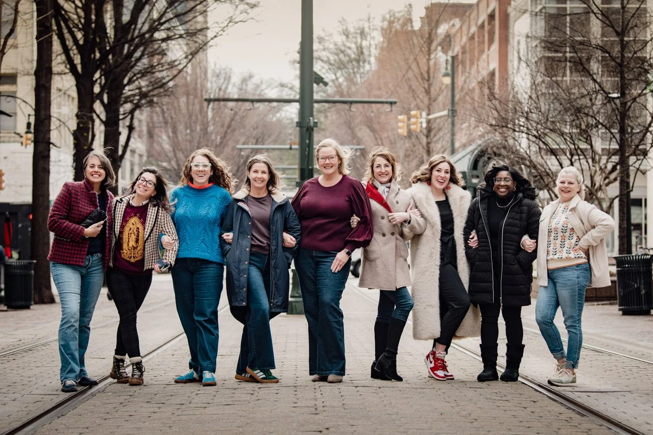 A group of ten women walking arm-in-arm on a city street, smiling and enjoying each other's company, with trees and buildings in the background.