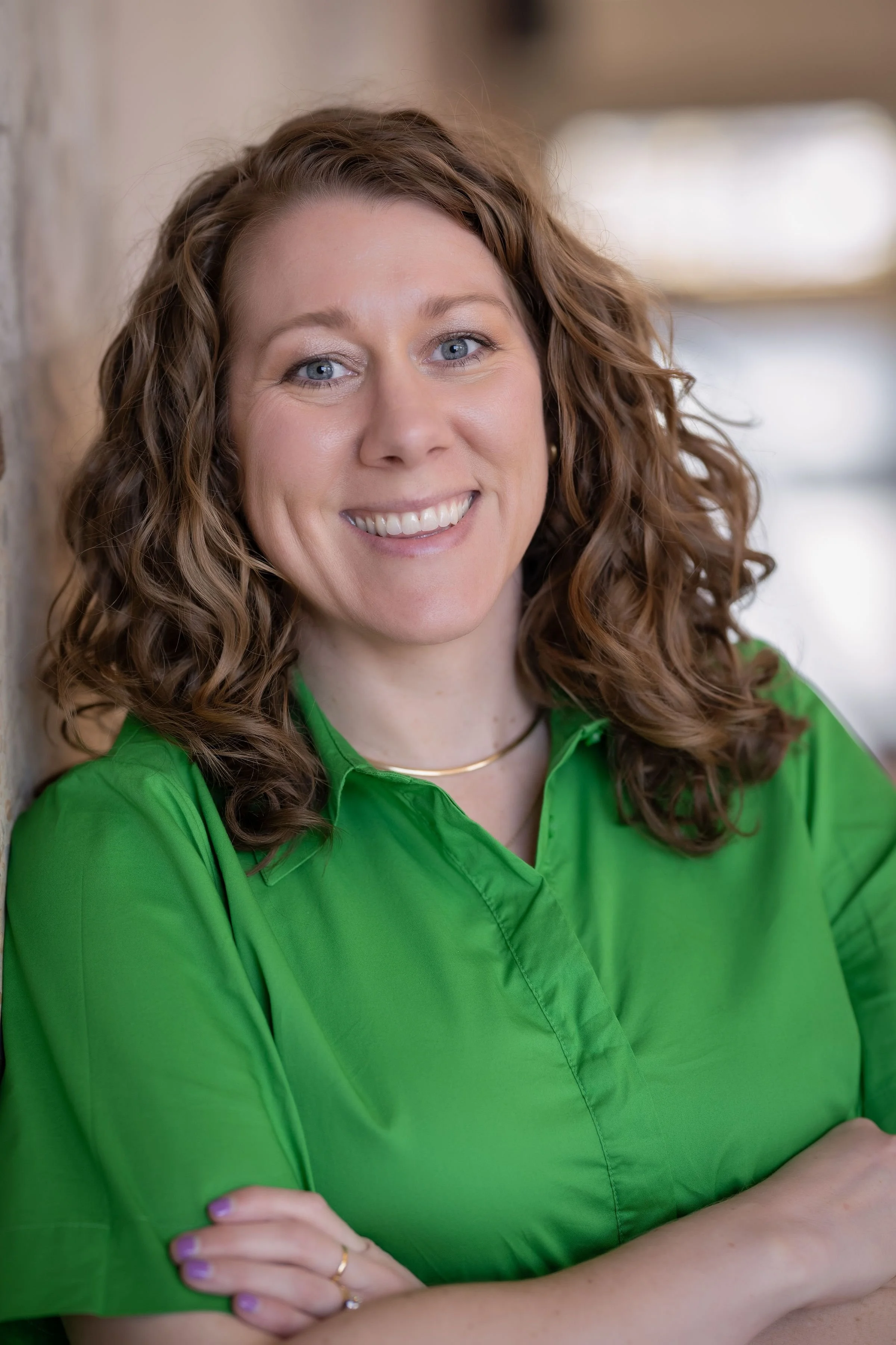 A woman with curly brown hair, wearing a green shirt, smiling with folded arms.