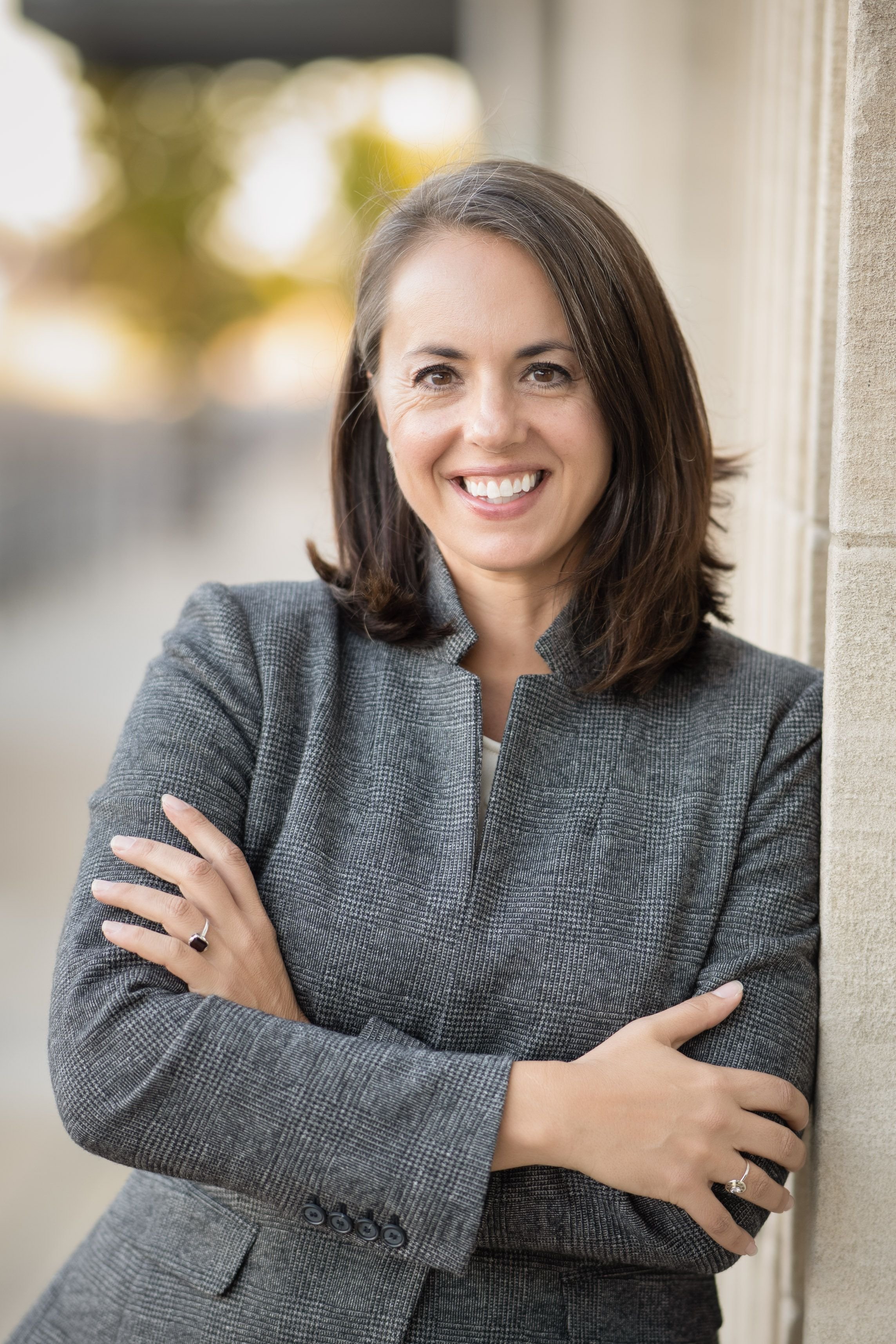 A smiling woman with shoulder-length brown hair wearing a gray plaid blazer, standing with arms crossed outdoors beside a wall with a blurred background of trees and sunlight.