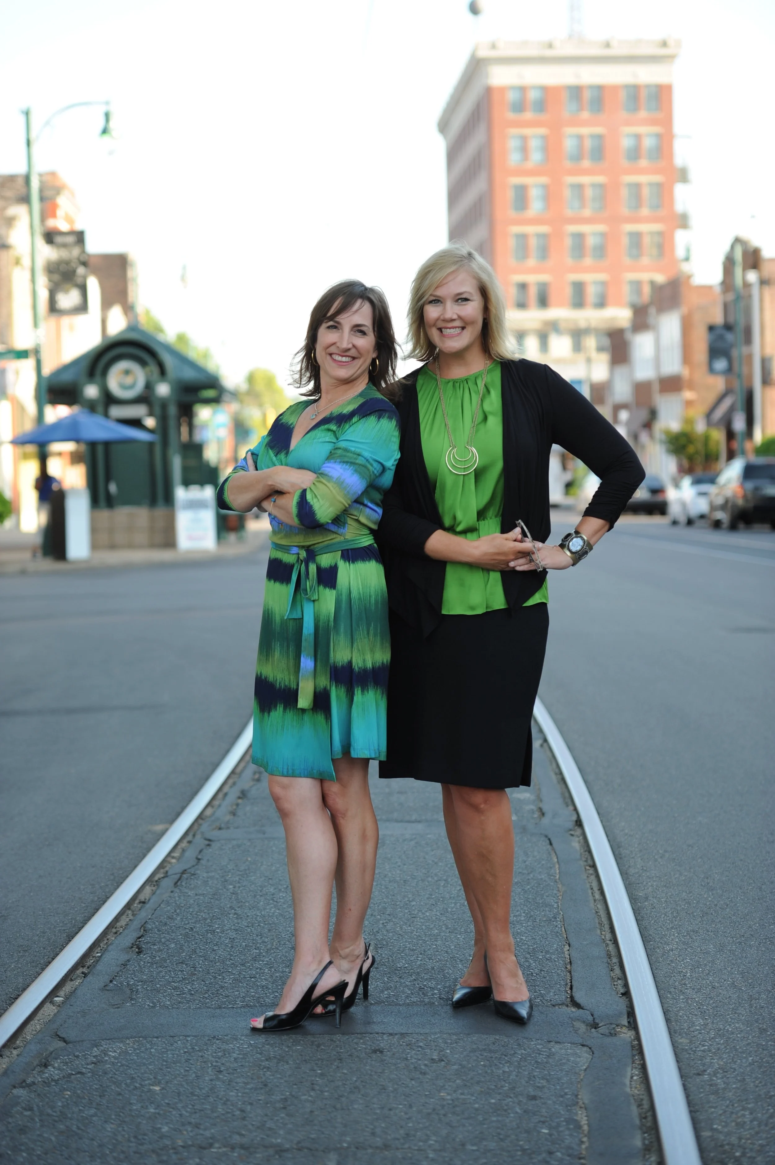 Two women standing on a street with a small train track, smiling, wearing green and black dresses, with buildings in the background.