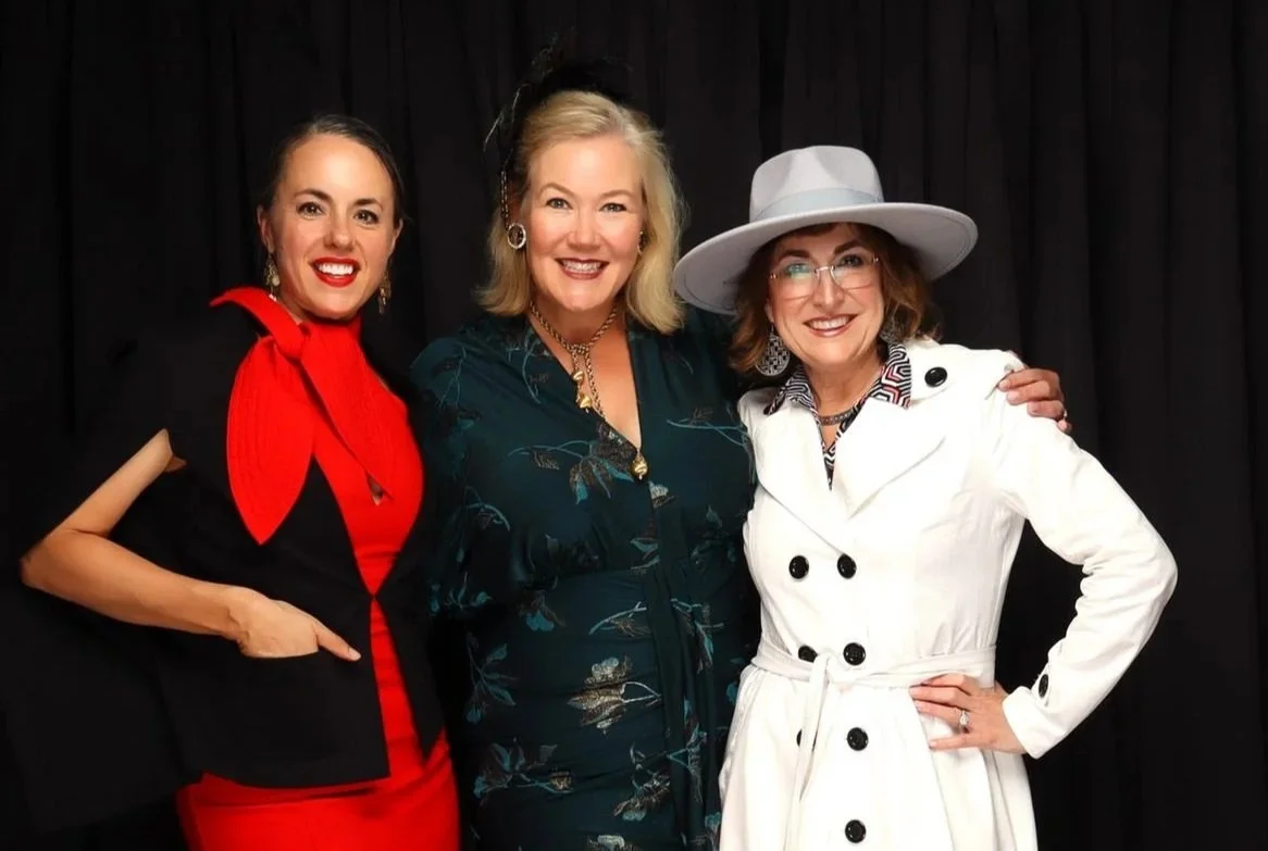 Andrea Wiley, Cynthia Saatkamp and Kelley Morice standing together in front of a black curtain, smiling at the camera, dressed in stylish outfits.