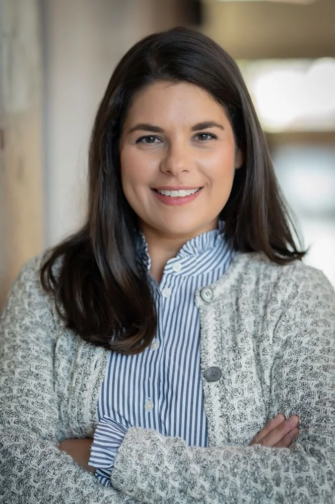 A woman with long dark brown hair smiling at the camera, wearing a blue and white striped button-up shirt underneath a light grey cardigan, with arms crossed.
